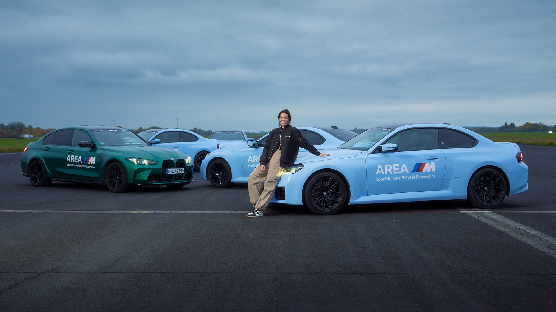 Eine Reihe von BMW M Fahrzeugen auf einer Asphaltbahn, mit einer grünen Limousine links und mehreren hellblauen Coupes. Die Fahrzeuge tragen das 'AREA M'-Branding, das auf BMWs Fahrerlebnisprogramm hinweist. Eine Person in schwarzer Jacke steht mit ausgestreckten Armen zwischen den Fahrzeugen unter einem bewölkten Himmel.
