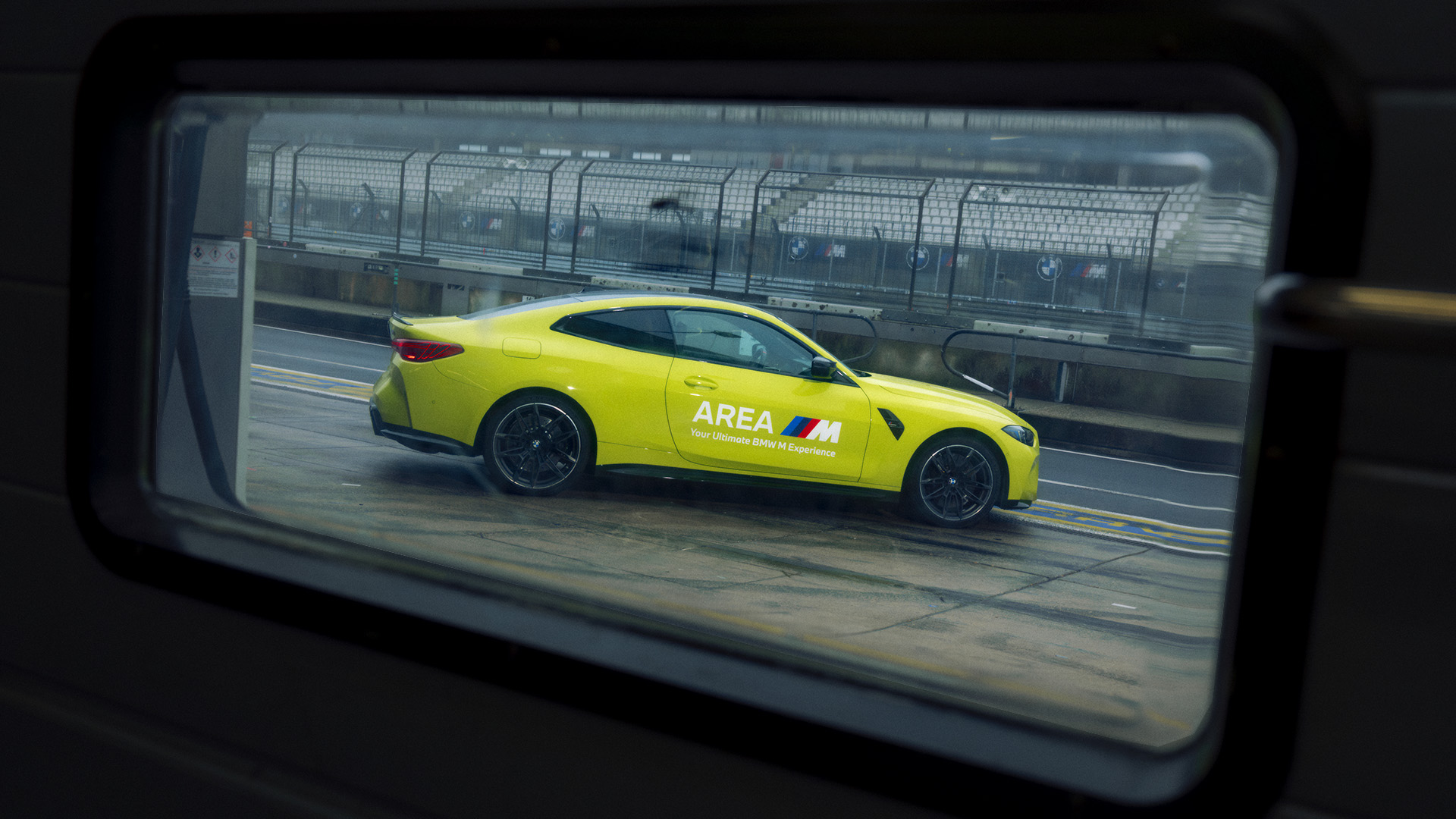 Ein leuchtend gelber BMW Sportwagen mit 'AREA M' Branding an der Seite, durch ein Fenster betrachtet auf was eine Rennstrecke zu sein scheint, mit Sicherheitszäunen im Hintergrund.