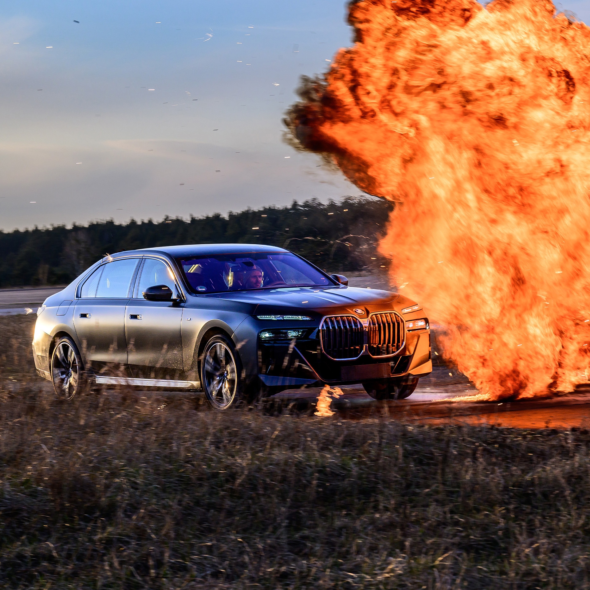 Ein schwarzer BMW Luxuswagen fährt auf einem Feld, während neben ihm eine große Explosion oder ein Feuer ausbricht. Die leuchtend orangefarbenen Flammen bilden einen dramatischen Kontrast zu dem dunklen Auto und der natürlichen Landschaft im Hintergrund.