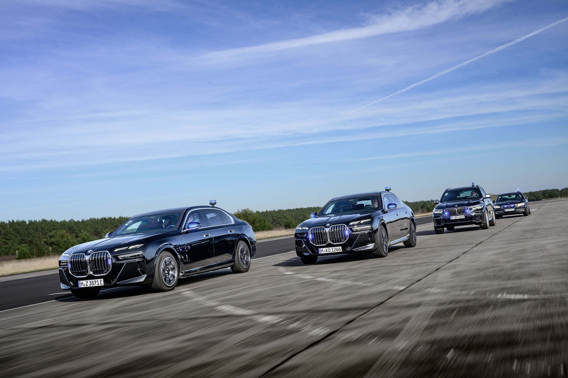 Vier schwarze BMW-Luxusfahrzeuge, die in Formation auf einer Teststrecke oder Landebahn fahren. Die Autos haben markante beleuchtete Nieren-Kühlergrills und werden in Bewegung vor einem Hintergrund aus klarem blauen Himmel mit dünnen Wolken und Kiefernwald in der Ferne fotografiert.