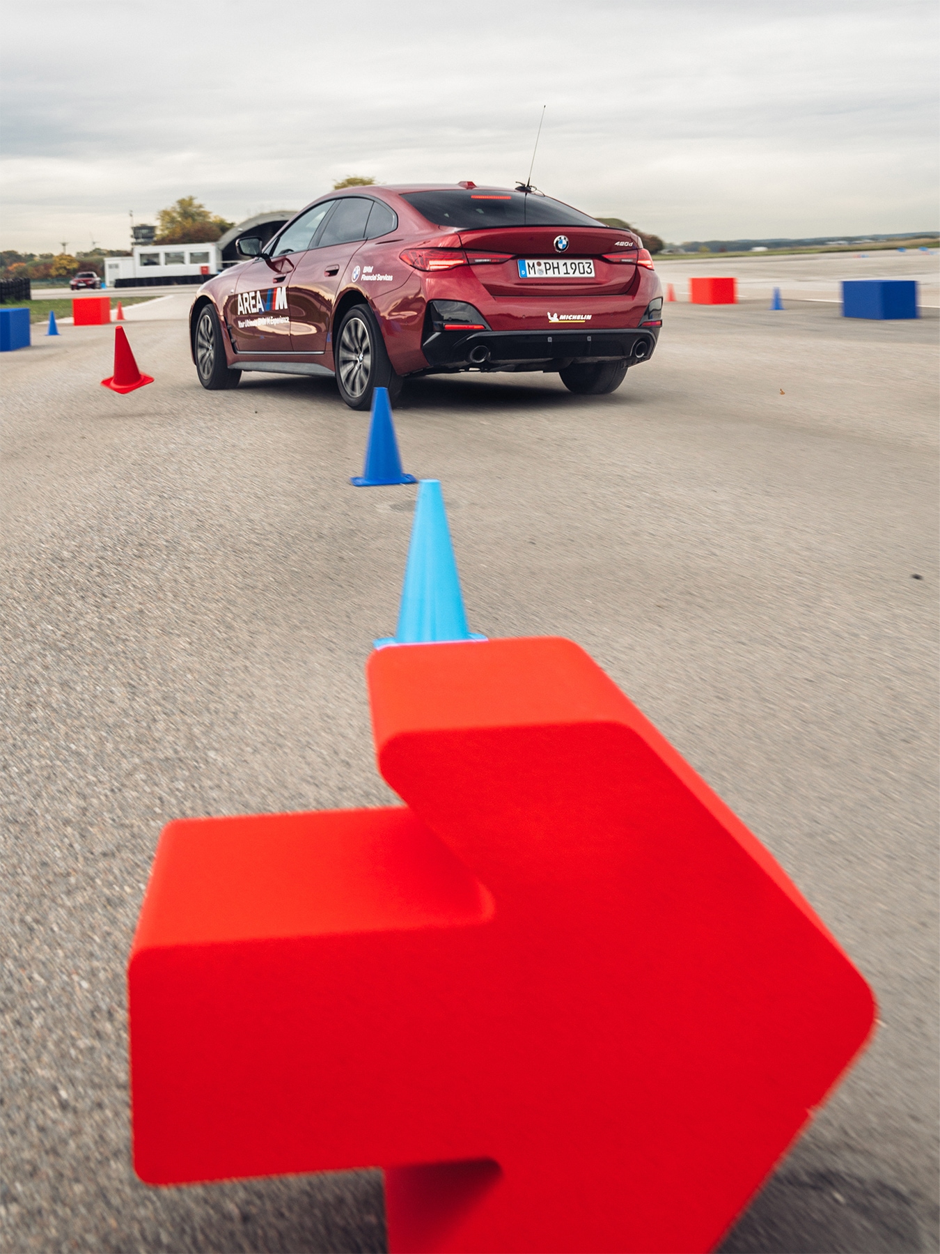 Ein burgunderroter BMW 4er Gran Coupe, der einen mit roten und blauen Verkehrskegeln markierten Fahrkurs auf einer asphaltierten Fläche durchfährt. Im Vordergrund ist ein roter Richtungspfeil zu sehen, und das Auto trägt die Aufschrift 'AREA M' an der Seite. Die Szene scheint auf einer Teststrecke oder Trainingsanlage unter bewölktem Himmel stattzufinden.