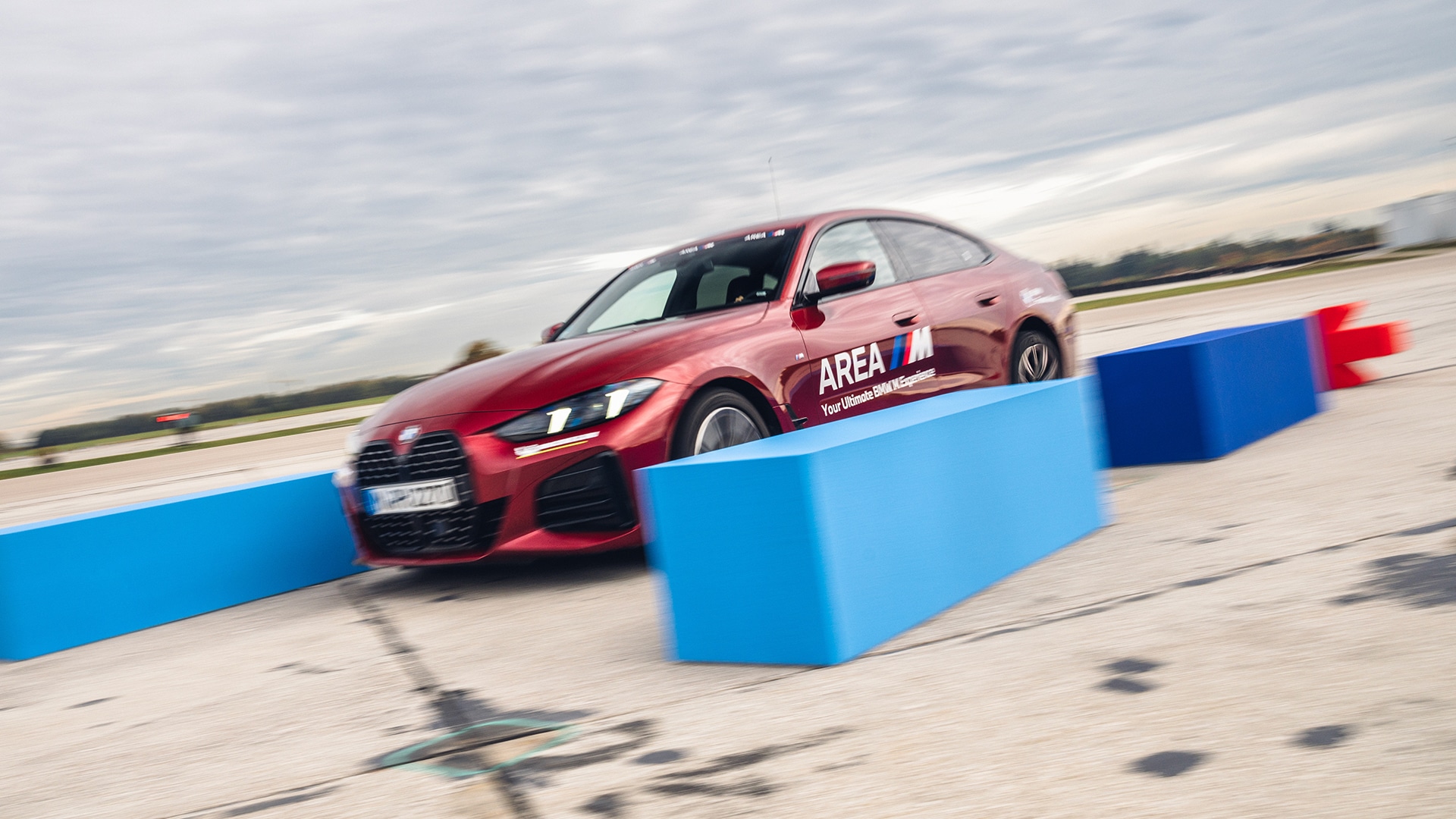 Ein roter BMW-Limousine mit 'AREA M'-Branding, der auf einem Fahrparcours zwischen blauen Barrieren unter bewölktem Himmel fährt.
