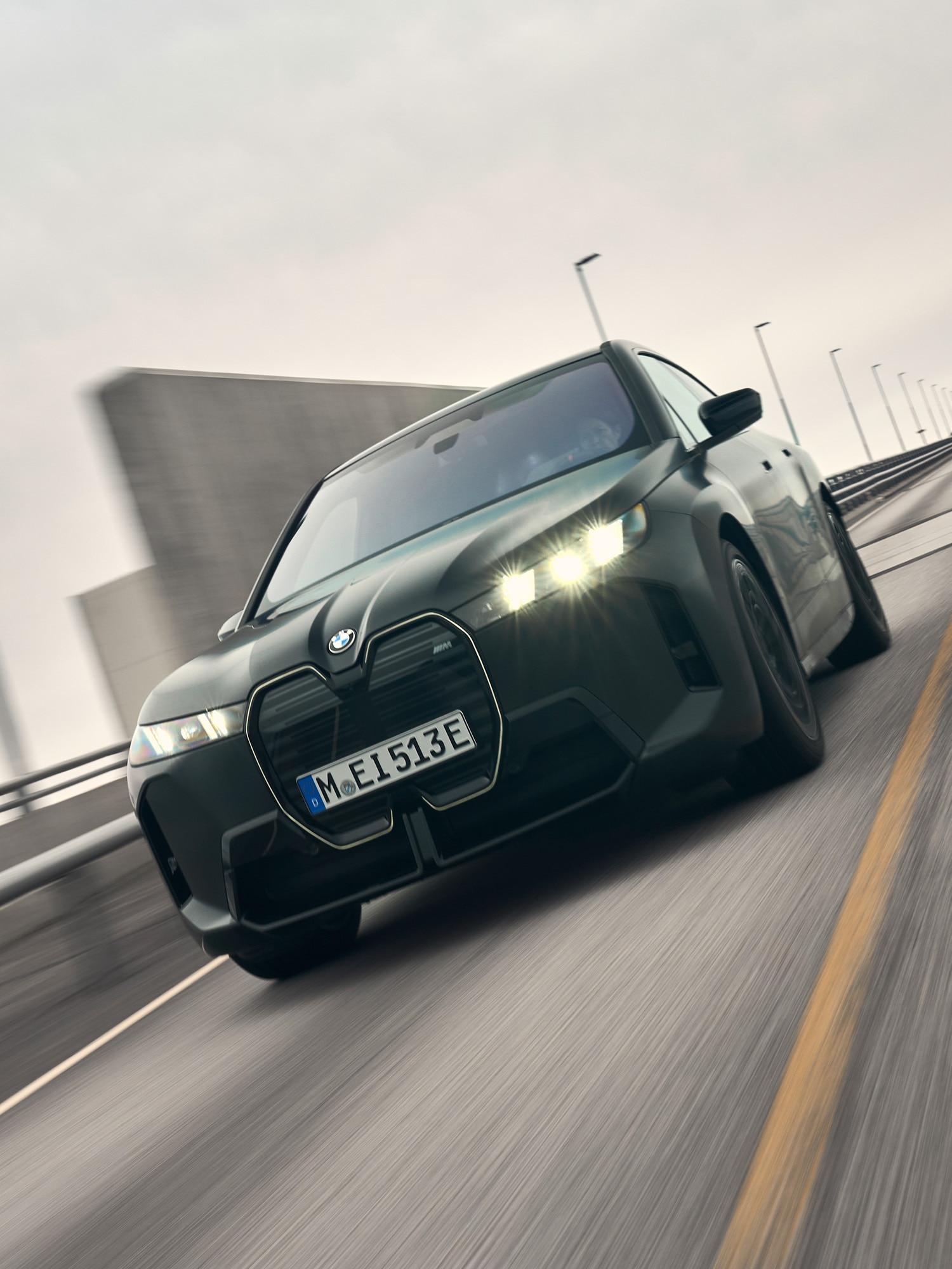 Ein schwarzes BMW Elektrofahrzeug mit dem Kennzeichen M-E 513E fährt auf einer Autobahn. Die Scheinwerfer des Autos leuchten vor einem bewölkten Himmel, und Straßenlaternen säumen die Straße. Der markante, große BMW-Nierengrill ist an der Frontseite des Fahrzeugs deutlich zu erkennen.