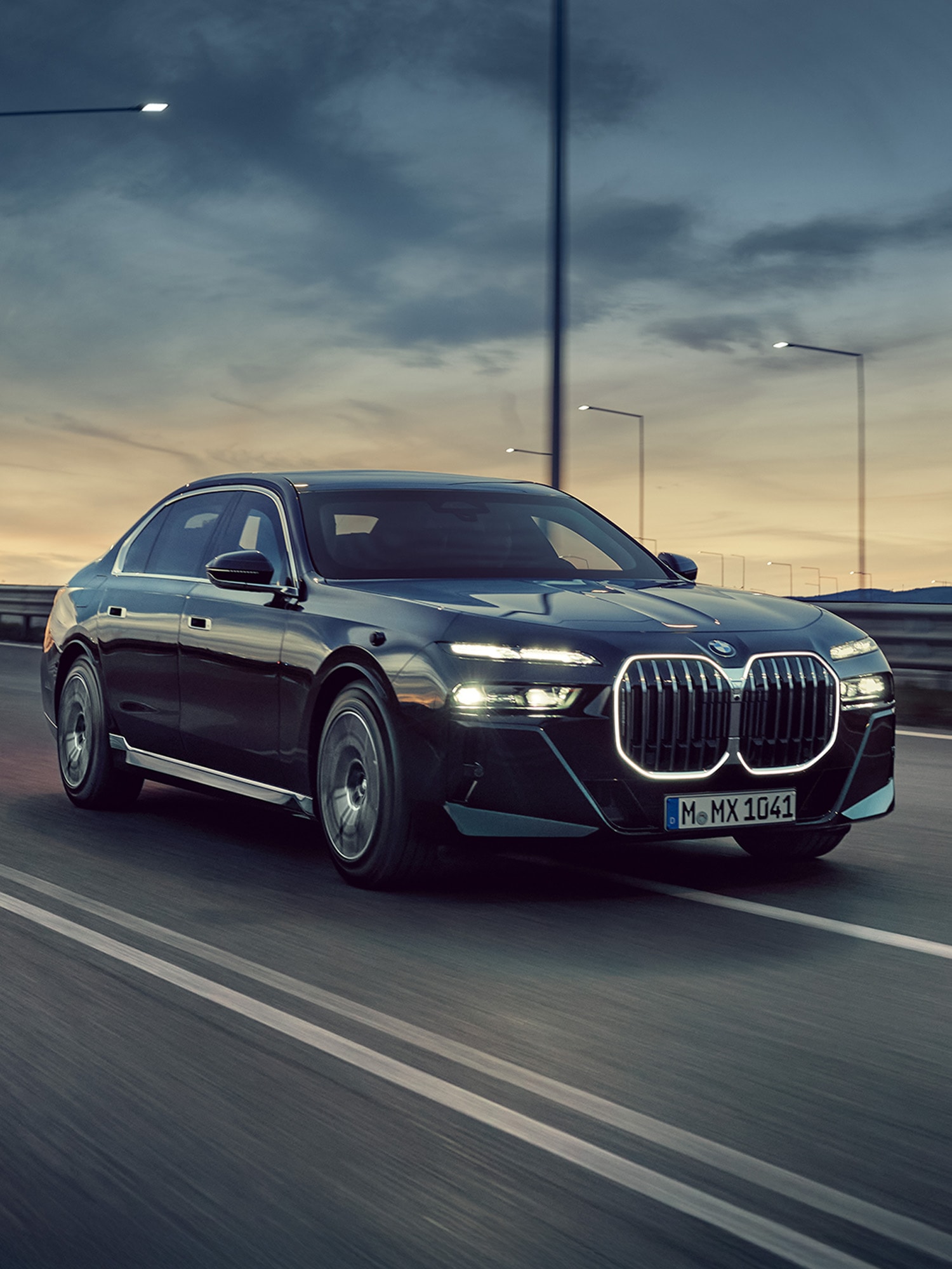 Ein schwarzer BMW Luxus-Sedan fährt bei Dämmerung auf einer Autobahn. Das Auto zeigt den markanten großen Kühlergrill und beleuchtete Scheinwerfer. Straßenlaternen säumen die Straße und der Himmel zeigt eine dramatische Mischung aus dunkelblau und goldenen Sonnenuntergangsfarben.