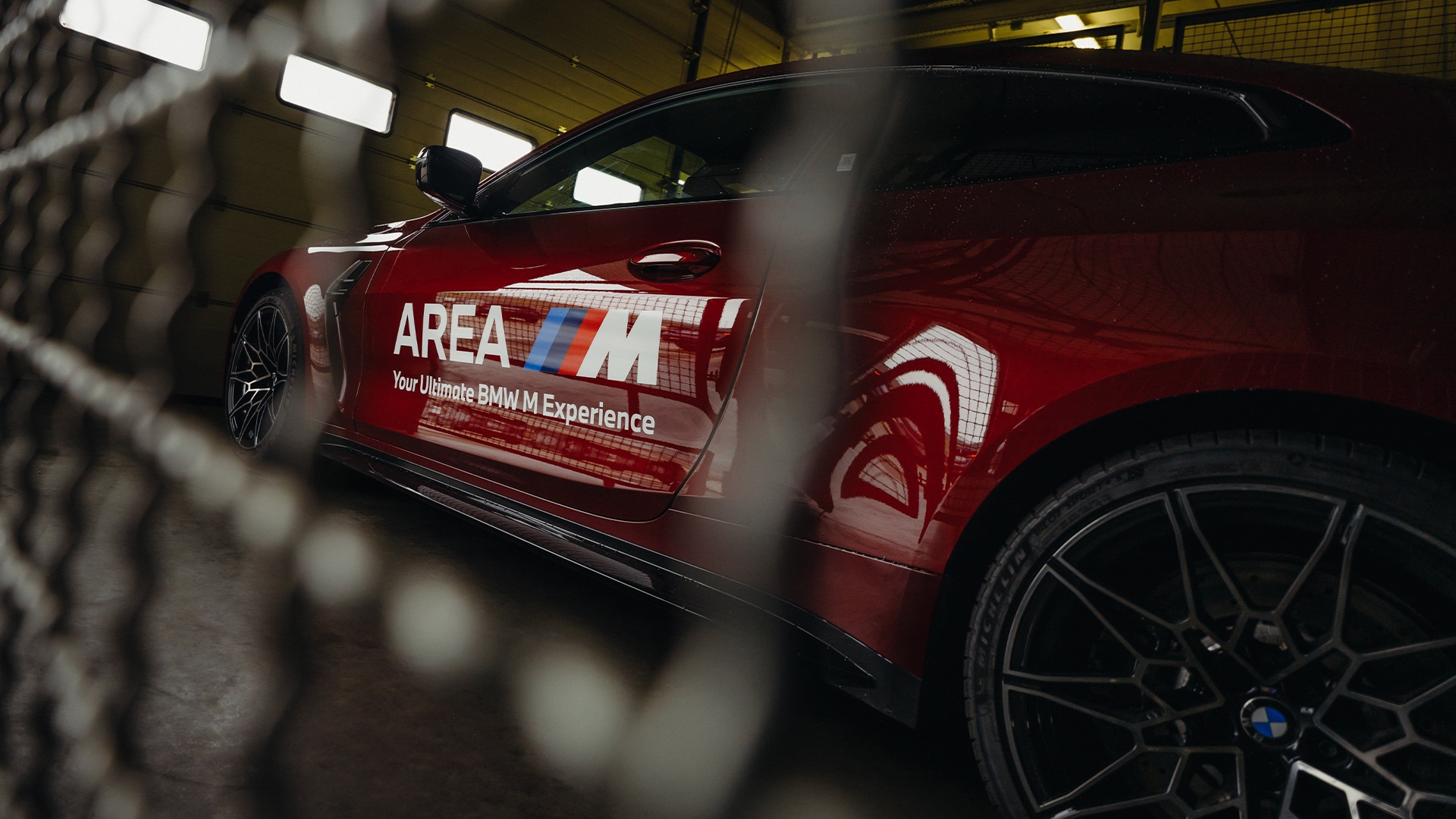 Ein glänzend roter BMW Sportwagen mit dem Text 'AREA M Your Ultimate BMW M Experience' auf der Tür, durch einen Maschendrahtzaun fotografiert. Das Fahrzeug steht in einer Halle mit Deckenbeleuchtung und zeigt sein elegantes Design und markante schwarze Leichtmetallfelgen.