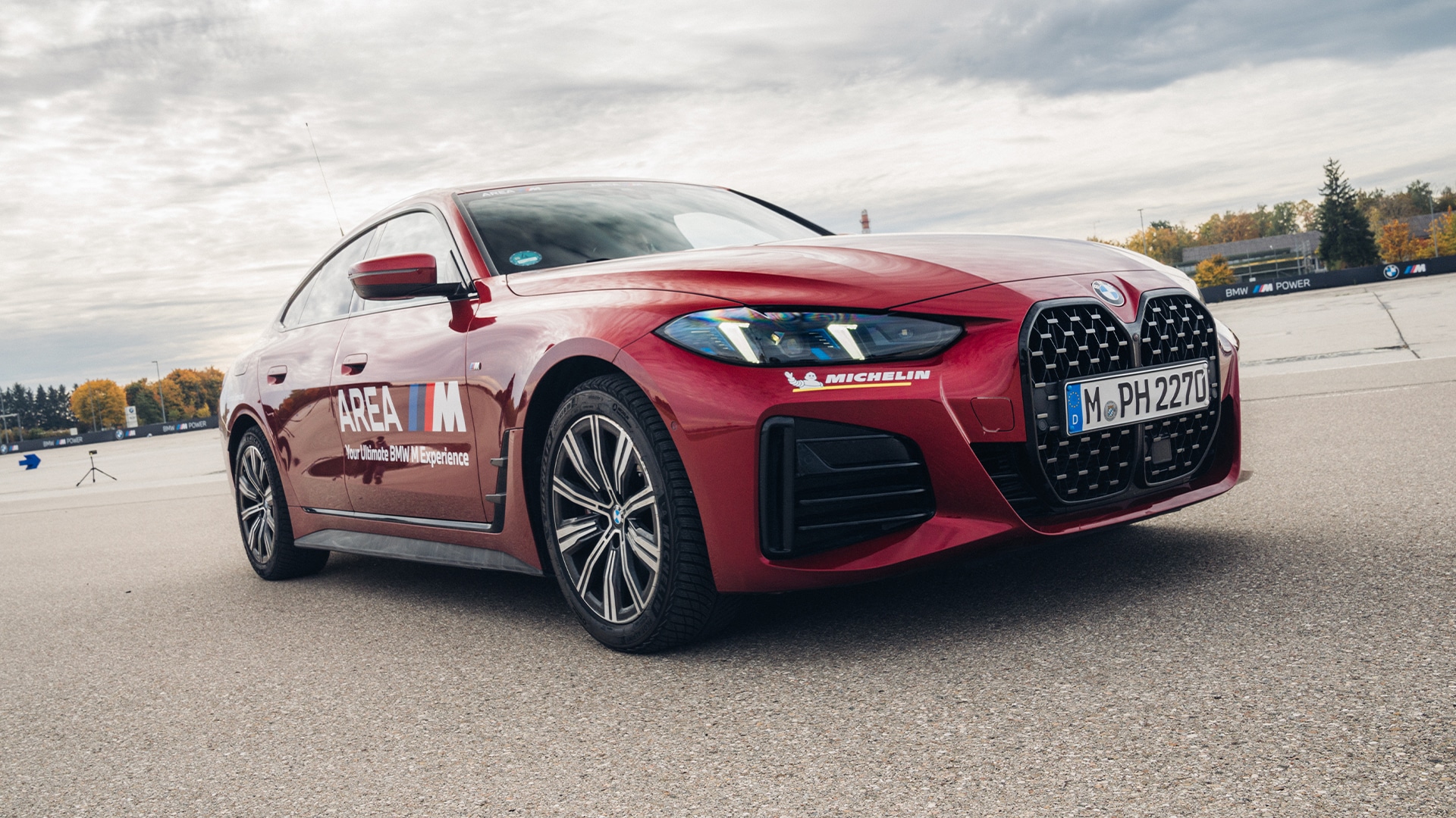 Ein roter BMW Sportwagen mit markanten großen Frontgrills auf einer Fahrstrecke geparkt. Das Fahrzeug zeigt 'AREA M' Branding an der Seite und hat ein Michelin-Logo an der Front. Die Szene zeigt einen bewölkten Himmel mit Herbstbäumen im Hintergrund.