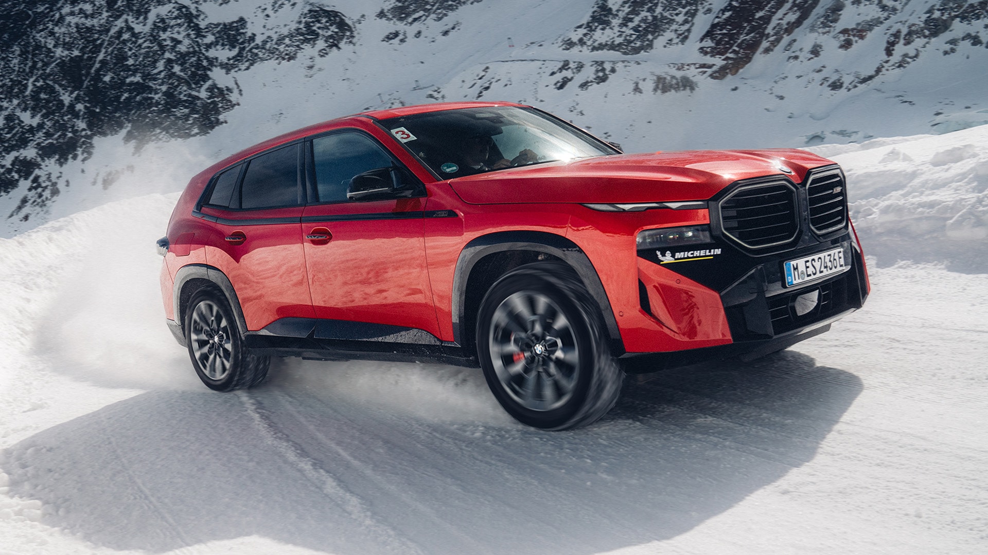 Ein leuchtend roter BMW SUV fährt durch eine verschneite Berglandschaft. Das Fahrzeug hat markante schwarze Räder und einen schwarzen Kühlergrill und wirbelt Schnee auf, während es sich über das weiße Gelände bewegt, mit schneebedeckten Bergen im Hintergrund.