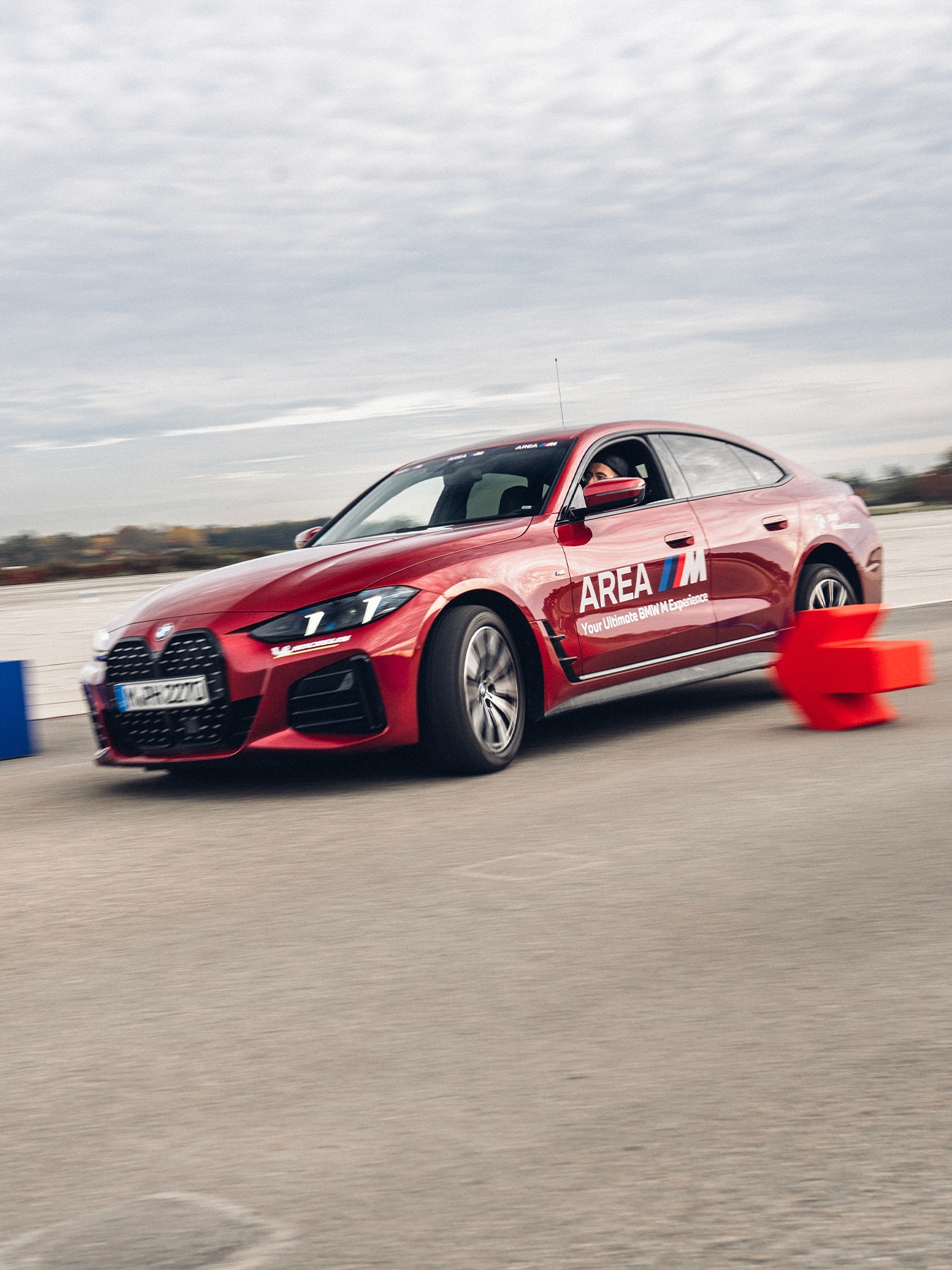Ein glänzender roter BMW Sedan mit "AREA M"-Branding auf der Seitentür ist auf einer Fahrtrainingsstrecke in Bewegung. Das Auto passiert orange Verkehrskegel auf einer asphaltierten Fläche unter einem bewölkten Himmel. Das Fahrzeug scheint Teil eines Fahrerlebnisses oder Trainingsprogramms zu sein.