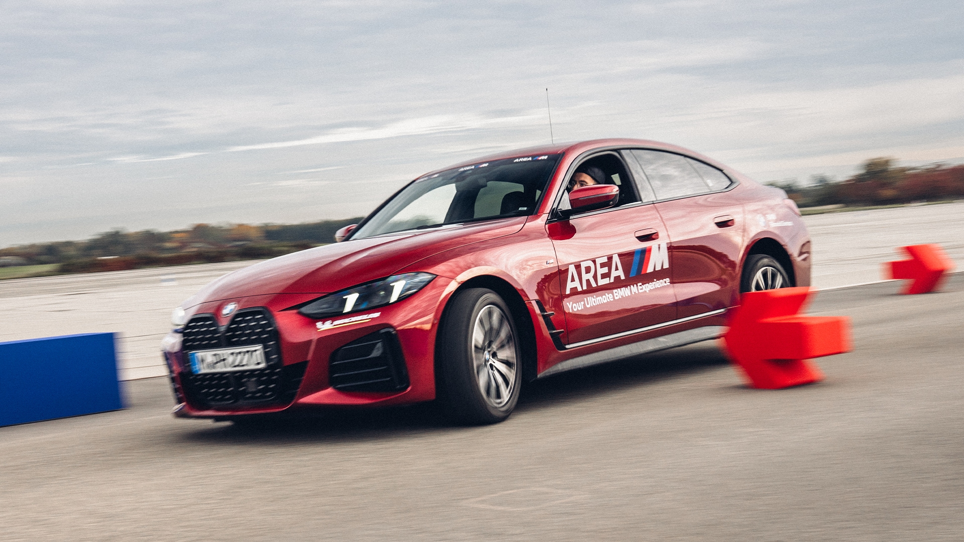 Ein roter BMW Sedan mit "AREA M" Beschriftung auf der Tür fährt auf einer Rennstrecke mit orangefarbenen Verkehrskegeln in Form eines Pfeils. Das Fahrzeug ist in Bewegung auf einer Betonfläche unter einem bewölkten Himmel.