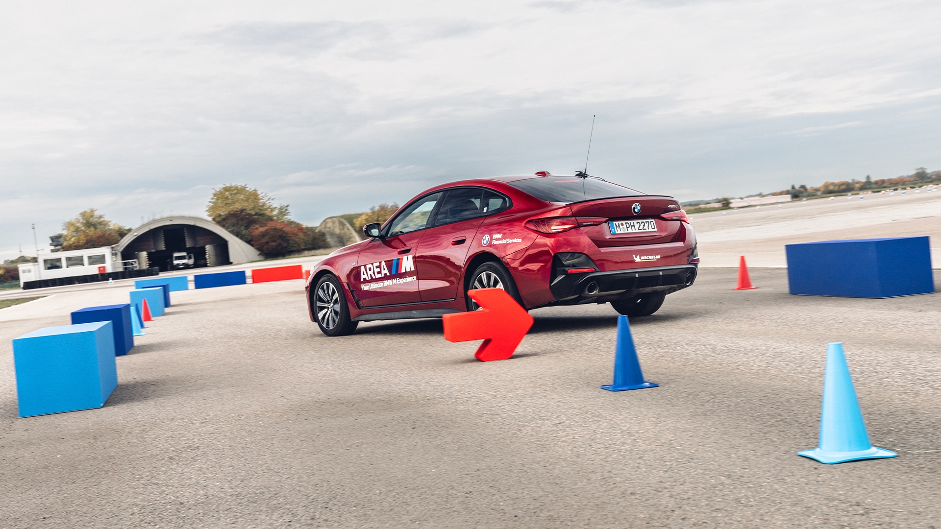 Ein roter BMW Sedan mit 'AREA M' Branding, der durch einen Fahrparcours mit blauen und roten Verkehrskegeln und Barrieren navigiert. Das Fahrzeug befindet sich auf einer Betonfläche, darüber ist ein bewölkter Himmel zu sehen und im Hintergrund sind einige Herbstbäume erkennbar.