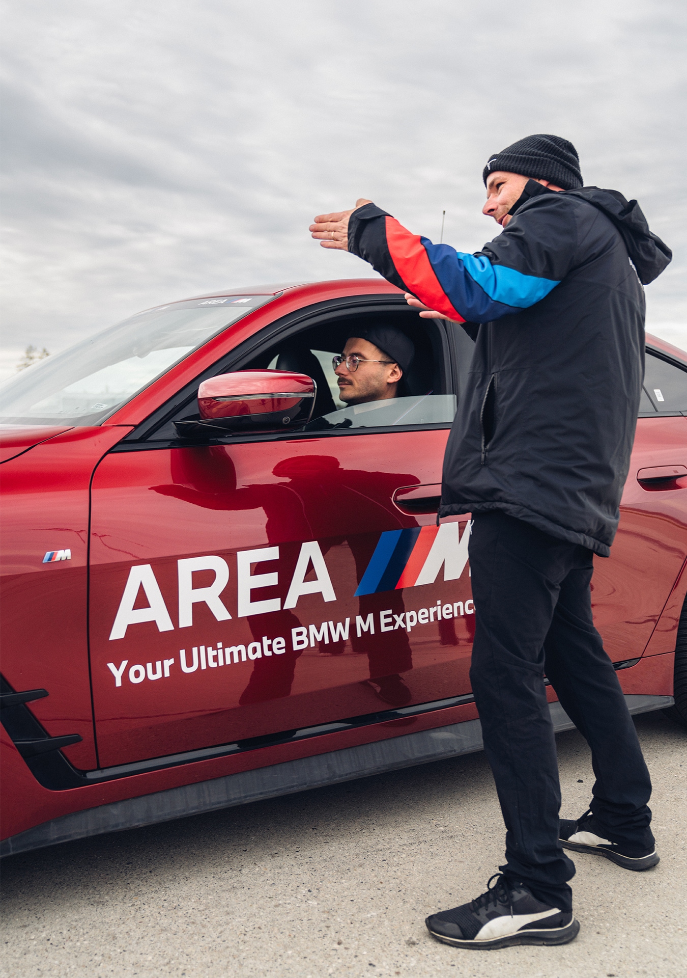 Eine Person in einer schwarzen Jacke mit bunten Ärmeln gibt jemandem Anweisungen, der in einem roten BMW Sportwagen sitzt. Auf der Autotür steht 'AREA M Your Ultimate BMW M Experience'.