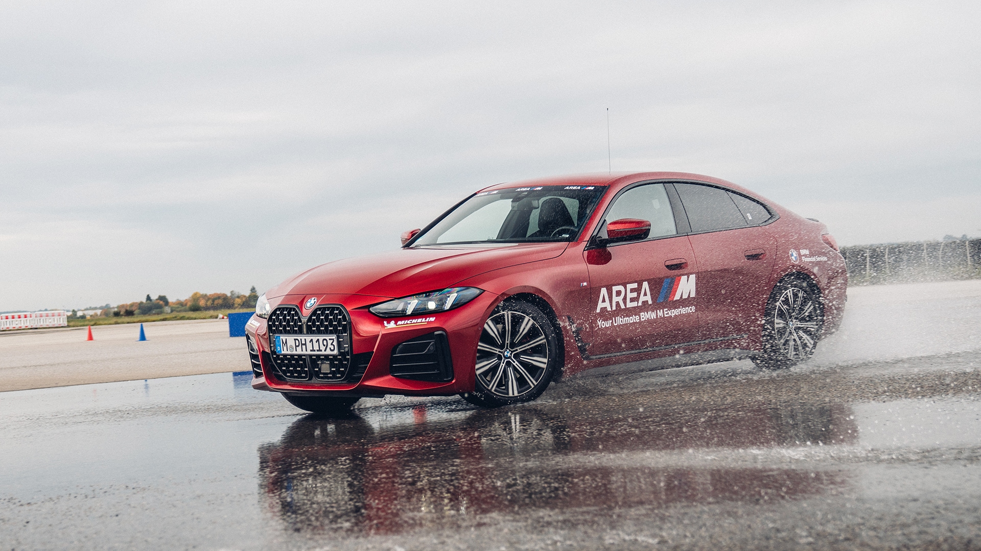 Ein roter BMW M Sedan fährt auf einer nassen Strecke, wobei Wasser von den Rädern spritzt. Das Fahrzeug trägt die Aufschrift 'AREA M' an der Seite und navigiert durch einen mit Verkehrskegeln markierten Kurs unter einem bewölkten Himmel.