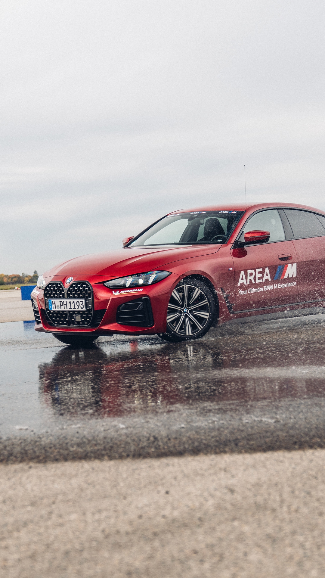Ein roter BMW Sportwagen mit der Aufschrift 'AREA M' auf der Seite, der auf einer nassen Fahrstrecke oder einem Kurs parkt. Die Oberfläche ist reflektierend mit Wasserpfützen, und der Himmel ist bewölkt. Das Fahrzeug zeigt das charakteristische BMW-Design mit dem markanten Kühlergrill und sportlichen Leichtmetallfelgen.