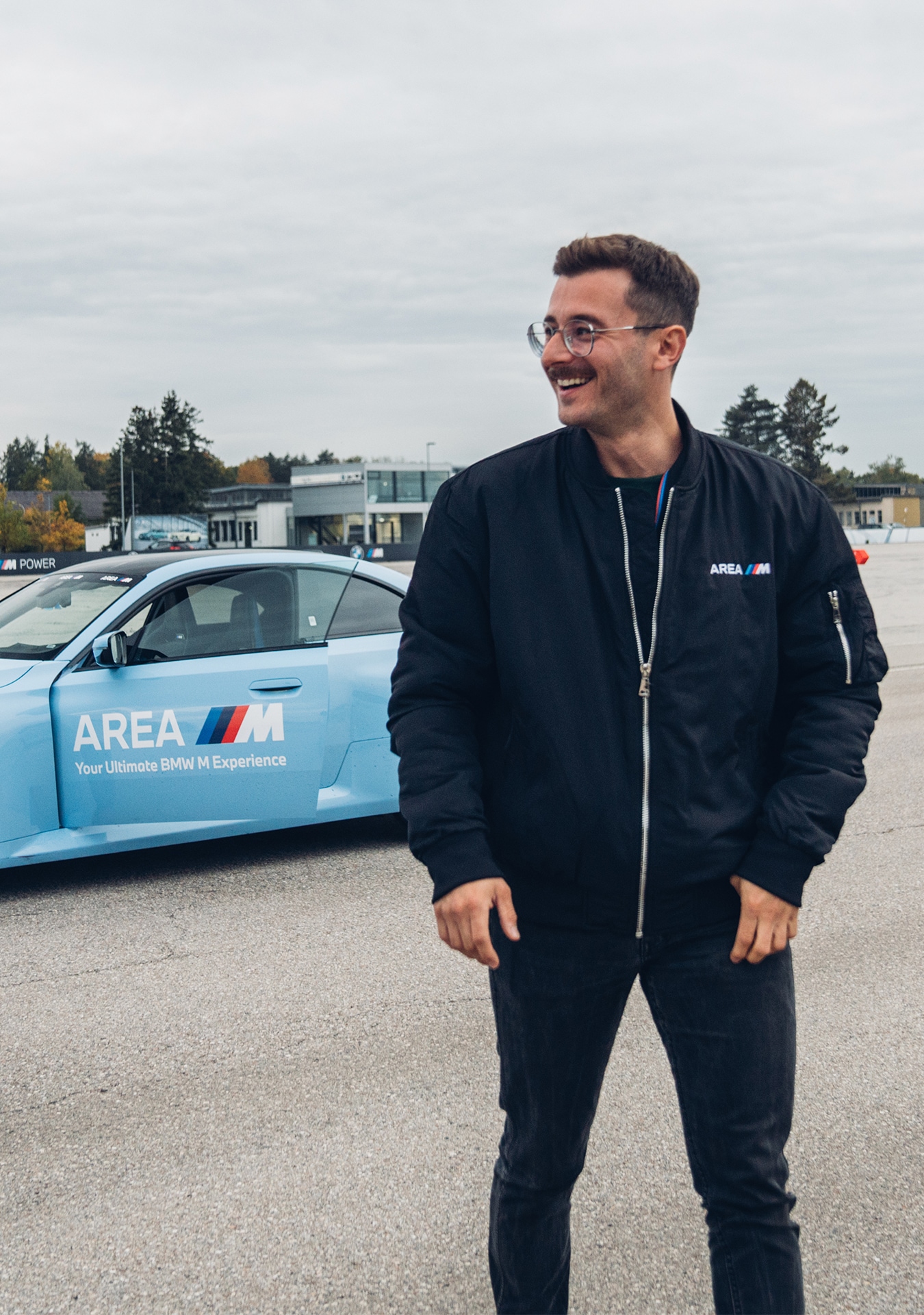 Eine Person mit Brille und schwarzer Bomberjacke mit AREA M Logo steht lächelnd auf einer gepflasterten Fläche. Hinter ihr steht ein hellblauer BMW, ebenfalls mit AREA M und dem Slogan 'Your Ultimate BMW M Experience' beschriftet. Im Hintergrund sind Gebäude und Bäume unter einem bewölkten Himmel zu sehen.