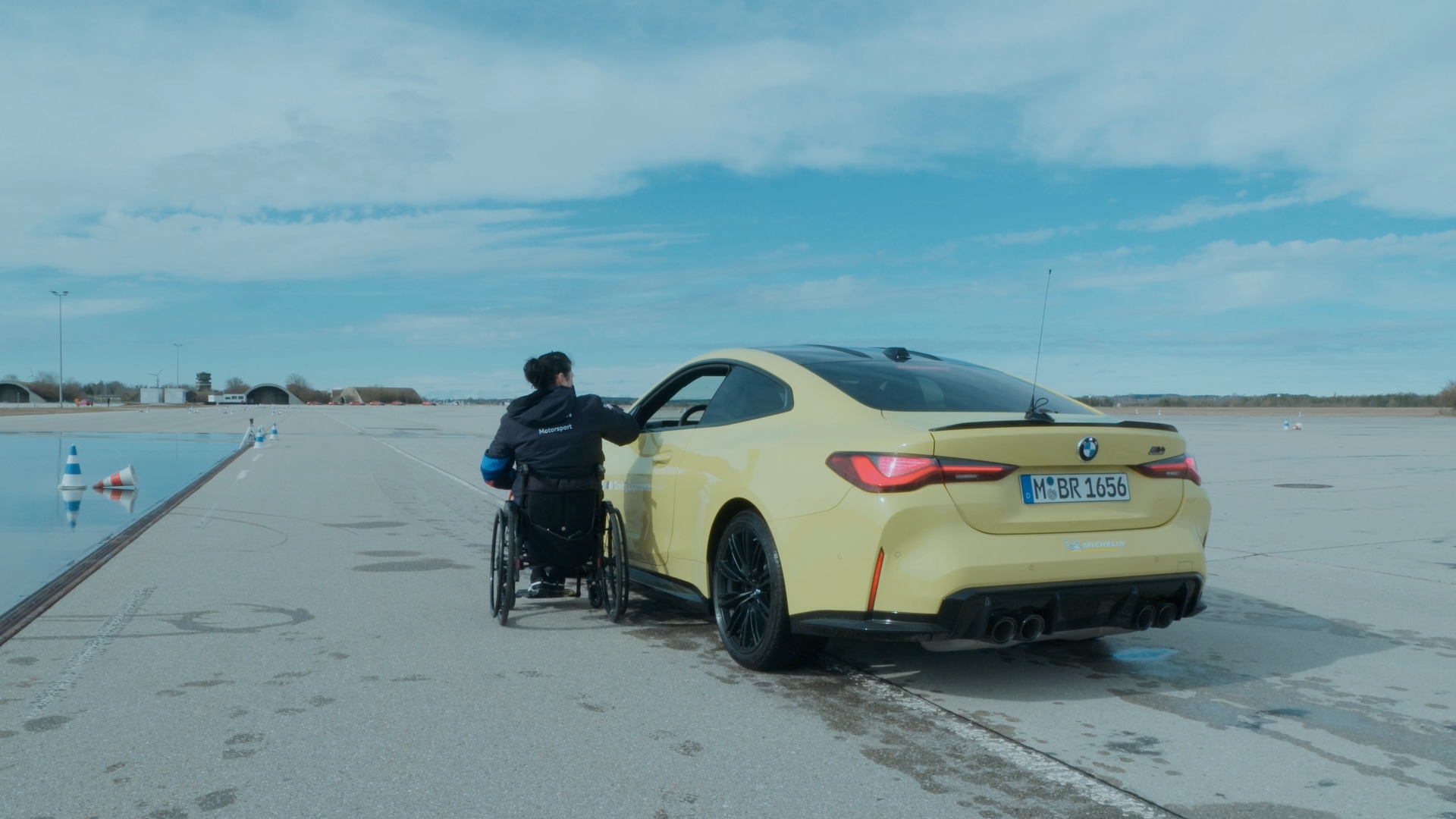Eine Person im Rollstuhl nähert sich der Fahrerseite eines gelben BMW M4 Sportwagens, der auf einer großen offenen Testfläche oder Strecke geparkt ist. Die Szene zeigt eine geräumige asphaltierte Fläche mit Verkehrskegeln in der Ferne unter einem blauen Himmel mit vereinzelten Wolken.