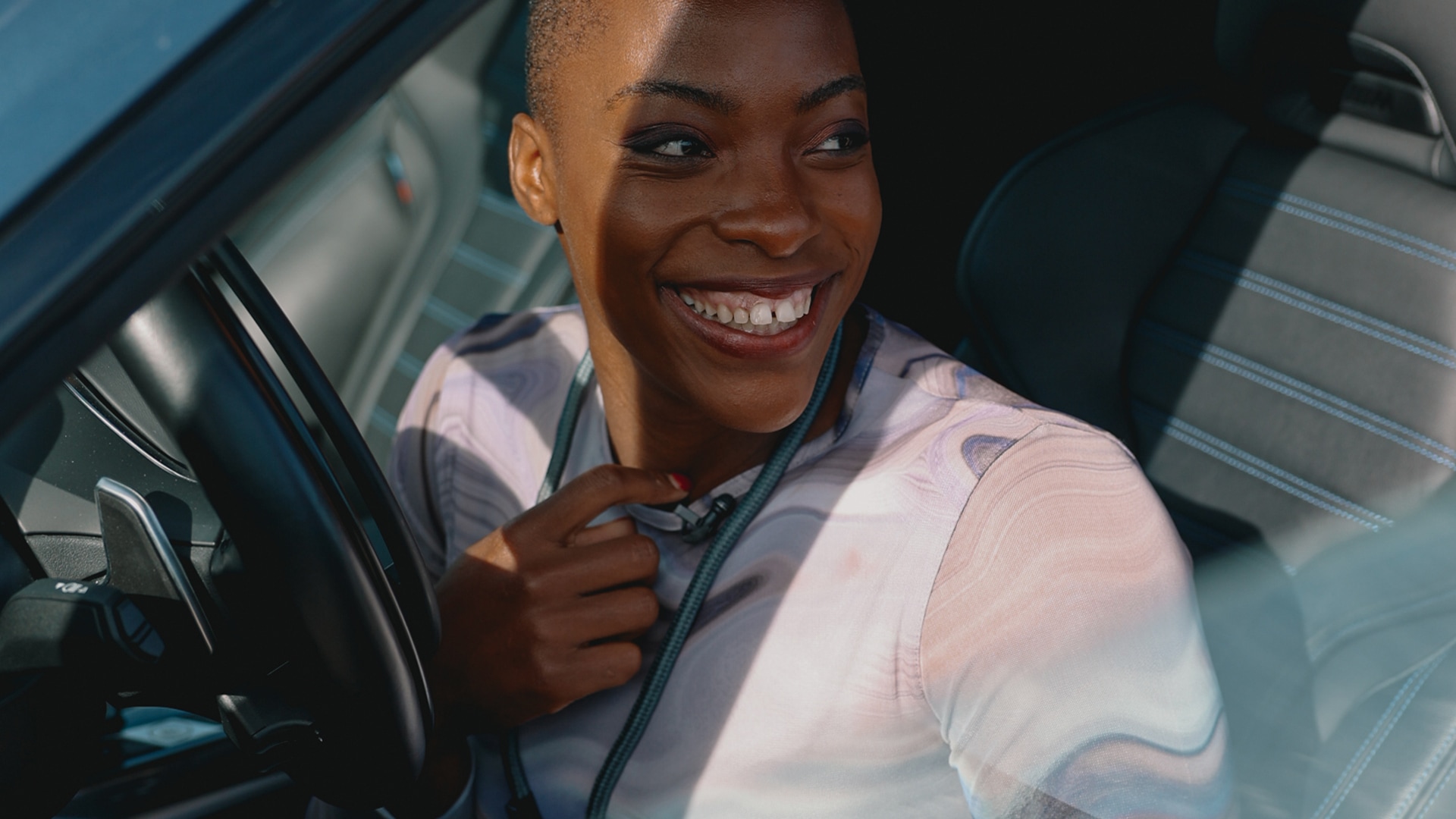 Eine Person mit kurzen Haaren sitzt auf dem Fahrersitz eines Autos und lächelt strahlend. Sie trägt ein helles, gemustertes Hemd. Im Innenraum des Fahrzeugs sind Teile des Lenkrads und der Sitzpolsterung mit sichtbaren Nähten zu erkennen.