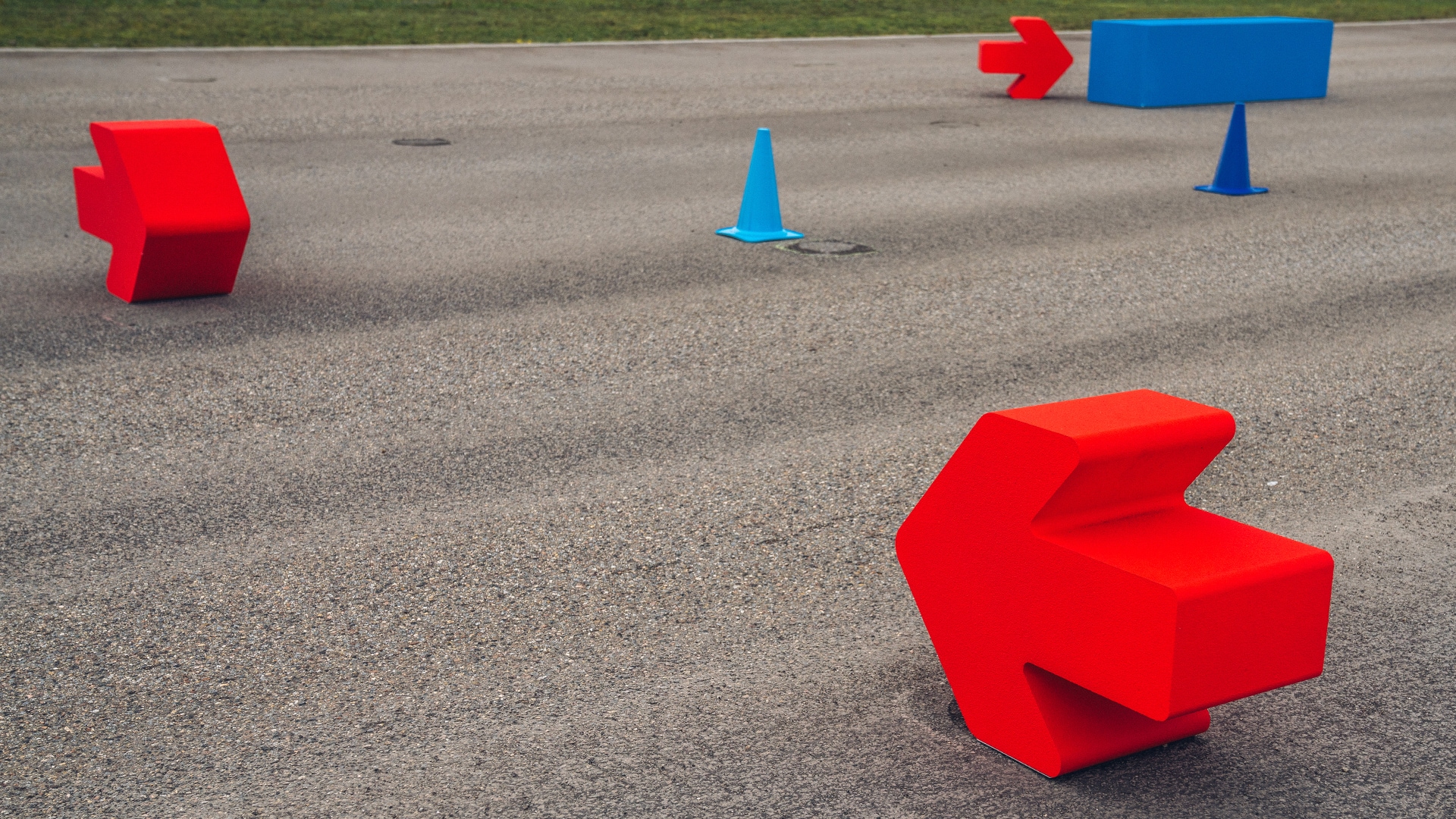 Eine gepflasterte Fläche mit Verkehrs- oder Trainingshindernissen, darunter rote pfeilförmige Blöcke, blaue Kegel und ein blaues rechteckiges Schild. Die roten und blauen Objekte scheinen Teil eines Fahrparcours oder einer Verkehrsmanagement-Einrichtung auf einer Asphaltfläche zu sein, wobei am oberen Rand Gras zu sehen ist.