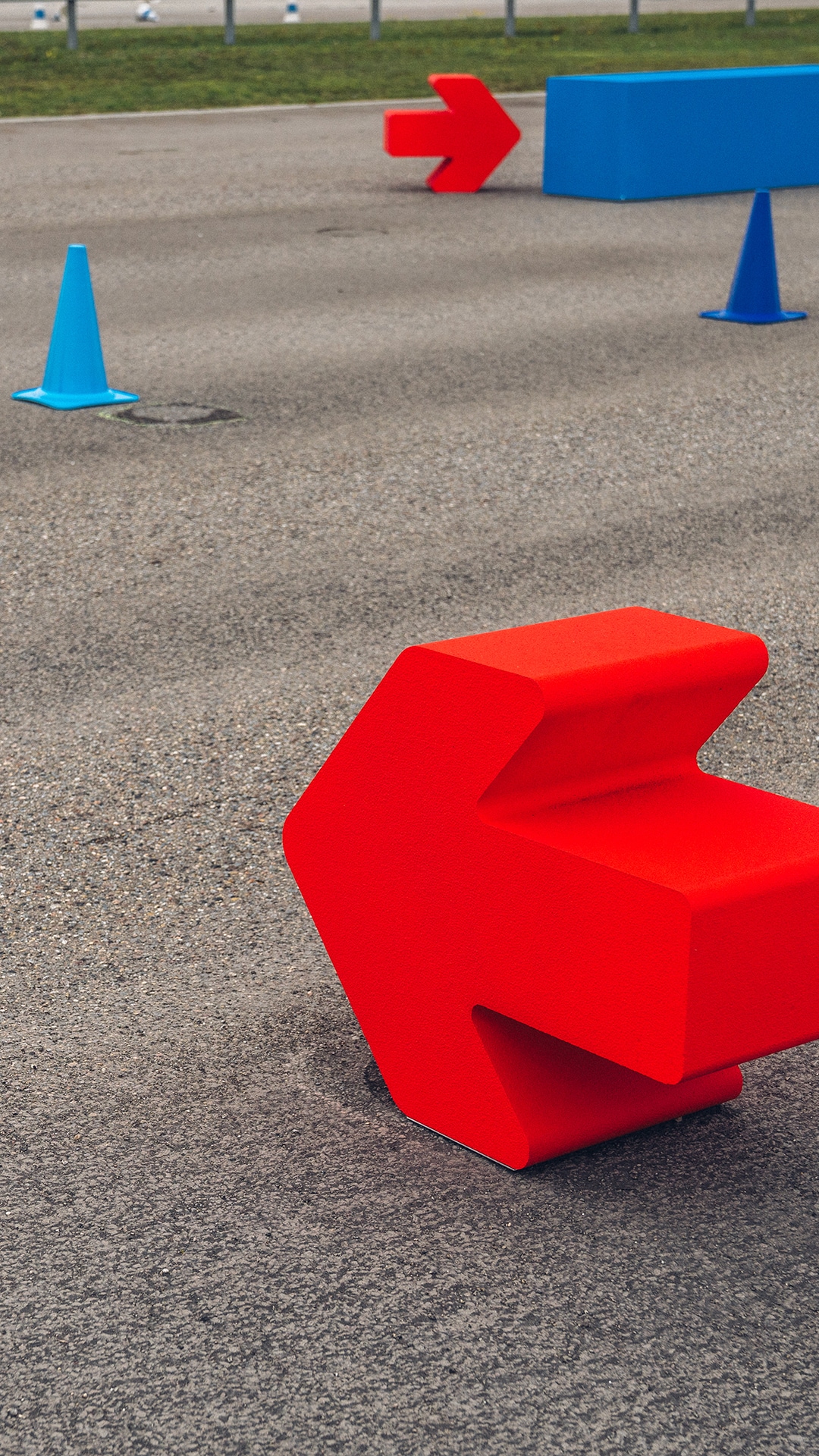 Ein Fahrparcours oder Trainingsgelände mit leuchtend roten pfeilförmigen Markierungen und blauen Verkehrskegeln auf einer Asphaltfläche. Im Vordergrund befindet sich eine große rote Richtungsmarkierung, im Hintergrund sind ein weiterer roter Pfeil und eine blaue Barriere zu sehen.