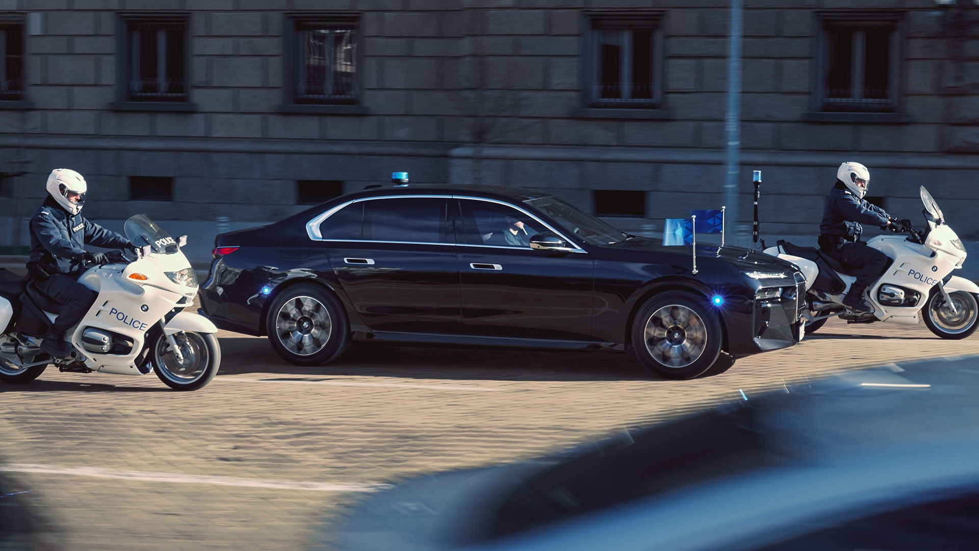 Eine schwarze Luxuslimousine mit offiziellen Flaggen und Blaulicht wird von zwei weißen Polizeimotorrädern eskortiert. Die Motorradfahrer tragen weiße Helme und dunkle Uniformen, während sie auf einer Kopfsteinpflasterstraße mit einem Steingebäude im Hintergrund fahren.