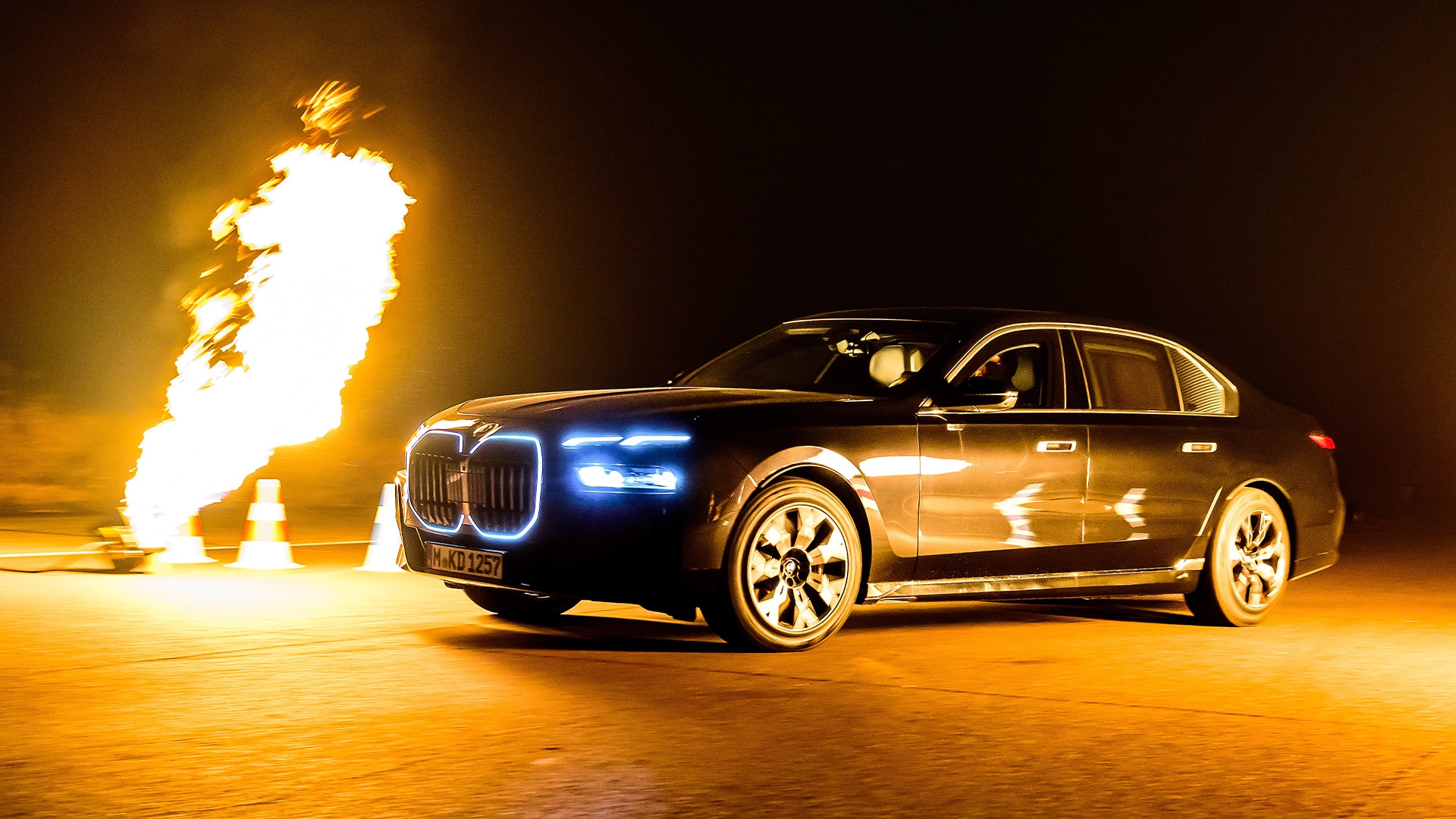 Eine schwarze BMW Luxuslimousine mit beleuchteten Nierengrillen und Scheinwerfern steht auf einer dunklen Oberfläche. Links schießt eine große Feuer- oder Flammensäule nach oben und erzeugt eine dramatische Beleuchtung vor dem nächtlichen Hintergrund.