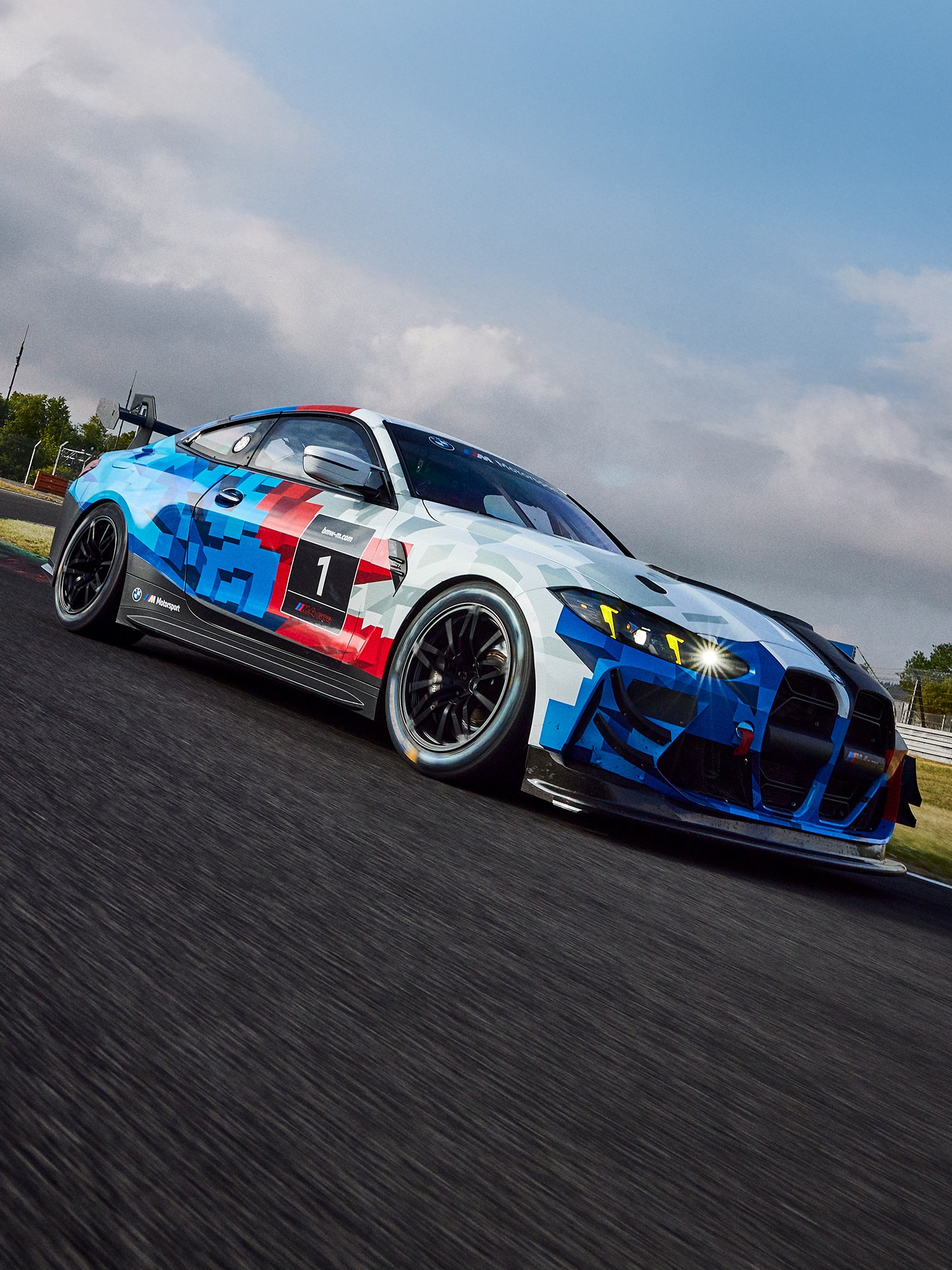Ein BMW-Rennwagen mit der Nummer 1 an der Seite, der eine markante geometrische Lackierung in Blau, Rot und Weiß aufweist. Das Hochleistungsfahrzeug ist auf einer Rennstrecke mit schwarzem Asphalt unter einem teilweise bewölkten Himmel fotografiert.