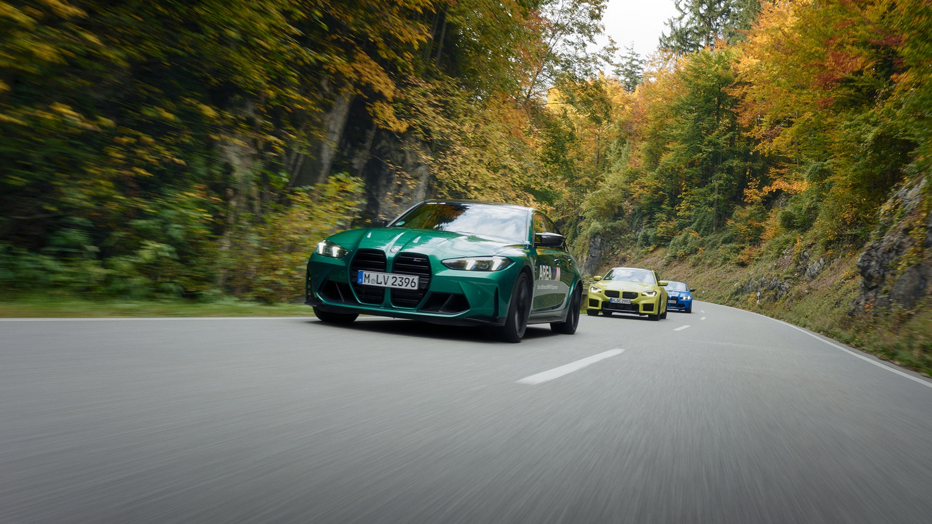 Ein grüner BMW Sportwagen führt zwei andere Fahrzeuge - ein gelbes und ein blaues Auto - auf einer kurvenreichen Bergstraße, umgeben von Herbstlaub in leuchtenden Orange-, Gelb- und Grüntönen. Die Straße schlängelt sich durch ein bewaldetes Gebiet mit sichtbaren Felswänden auf einer Seite.