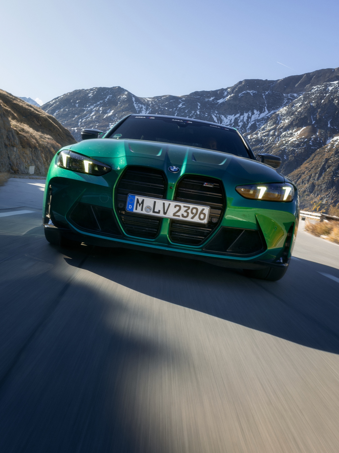 Ein metallisch grüner BMW Sportwagen mit markanten großen Nierengrillen fährt auf einer Bergstraße. Das Fahrzeug hat ein deutsches Kennzeichen und wird von vorne vor einem Hintergrund schneebedeckter Berge unter klarem blauen Himmel fotografiert.
