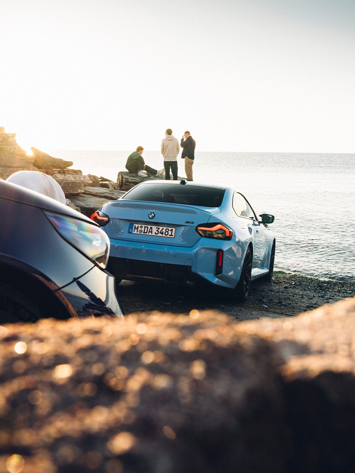 Ein hellblauer BMW M2 Sportwagen, der bei Sonnenuntergang am Ufer geparkt ist. Drei Personen stehen auf felsigen Vorsprüngen in der Nähe des Wassers und genießen den Meeresblick. Das deutsche Kennzeichen des Autos ist sichtbar, und goldenes Sonnenlicht spiegelt sich im Wasser und auf den umliegenden Felsen wider.