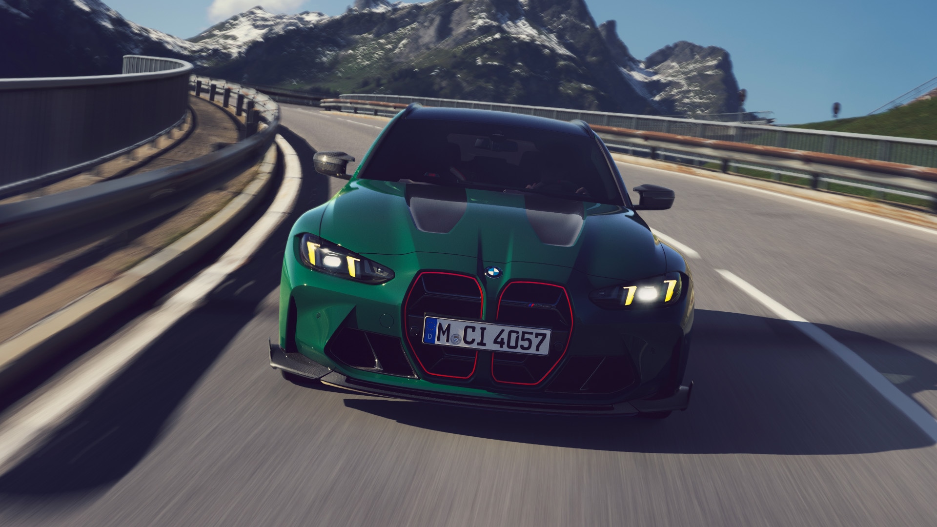 Ein grüner BMW Sportwagen mit markanten rot umrandeten Nieren fährt auf einer kurvenreichen Bergstraße. Das Fahrzeug mit dem Kennzeichen M-CI 4057 hat die Scheinwerfer eingeschaltet und bewegt sich auf einer malerischen Strecke mit schneebedeckten Bergen im Hintergrund vor einem klaren blauen Himmel.