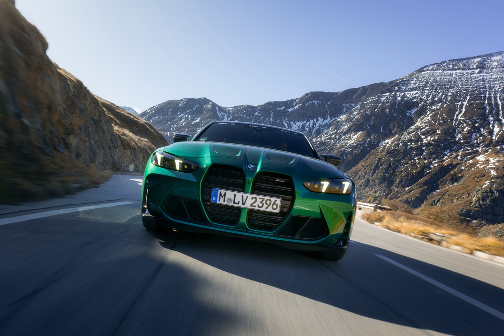 Ein metallisch grüner BMW Sportwagen fährt auf einer Bergstraße. Das Fahrzeug zeigt den markanten großen BMW-Nierengrill und ist von vorne während der Fahrt fotografiert. Im Hintergrund sind beeindruckende, leicht schneebedeckte Berge unter einem klaren blauen Himmel zu sehen.