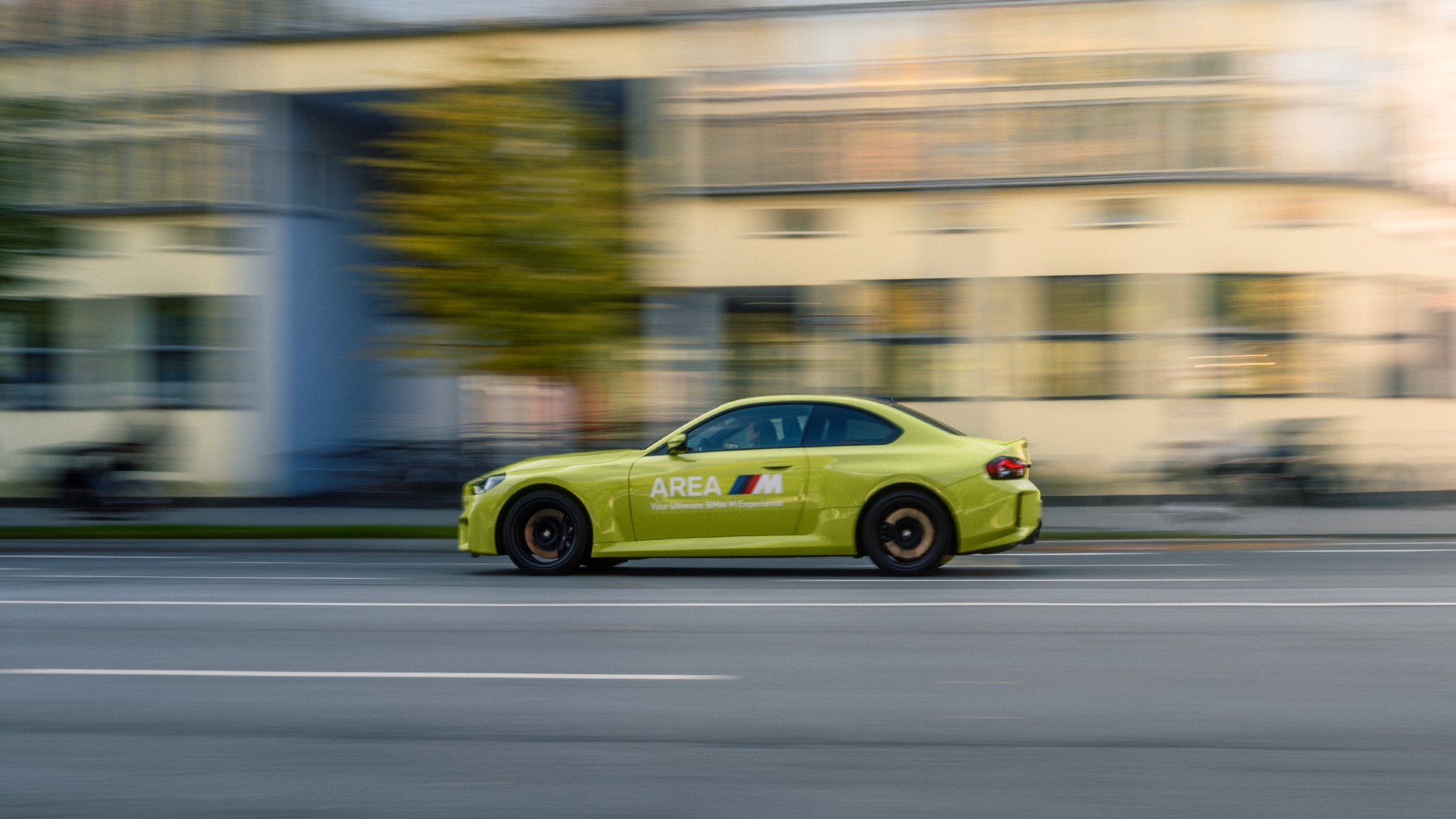 Ein leuchtend gelbes Sportcoupé mit 'AREA M'-Branding an der Seite, das mit hoher Geschwindigkeit auf einer Straße fährt. Der Hintergrund ist verschwommen und zeigt Gebäude und einen Baum, was den Eindruck von Bewegung vermittelt.