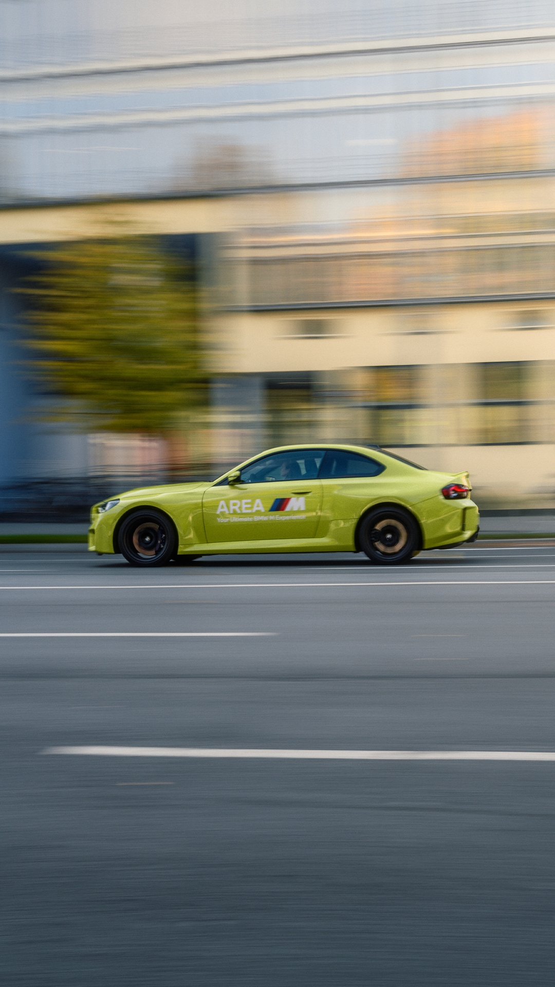 Ein leuchtend gelbgrüner Sportwagen mit 'AREA M'-Branding an der Seite, in Bewegung auf einer Straße. Der Hintergrund ist aufgrund der Geschwindigkeit verschwommen und zeigt ein Gebäude und Bäume.