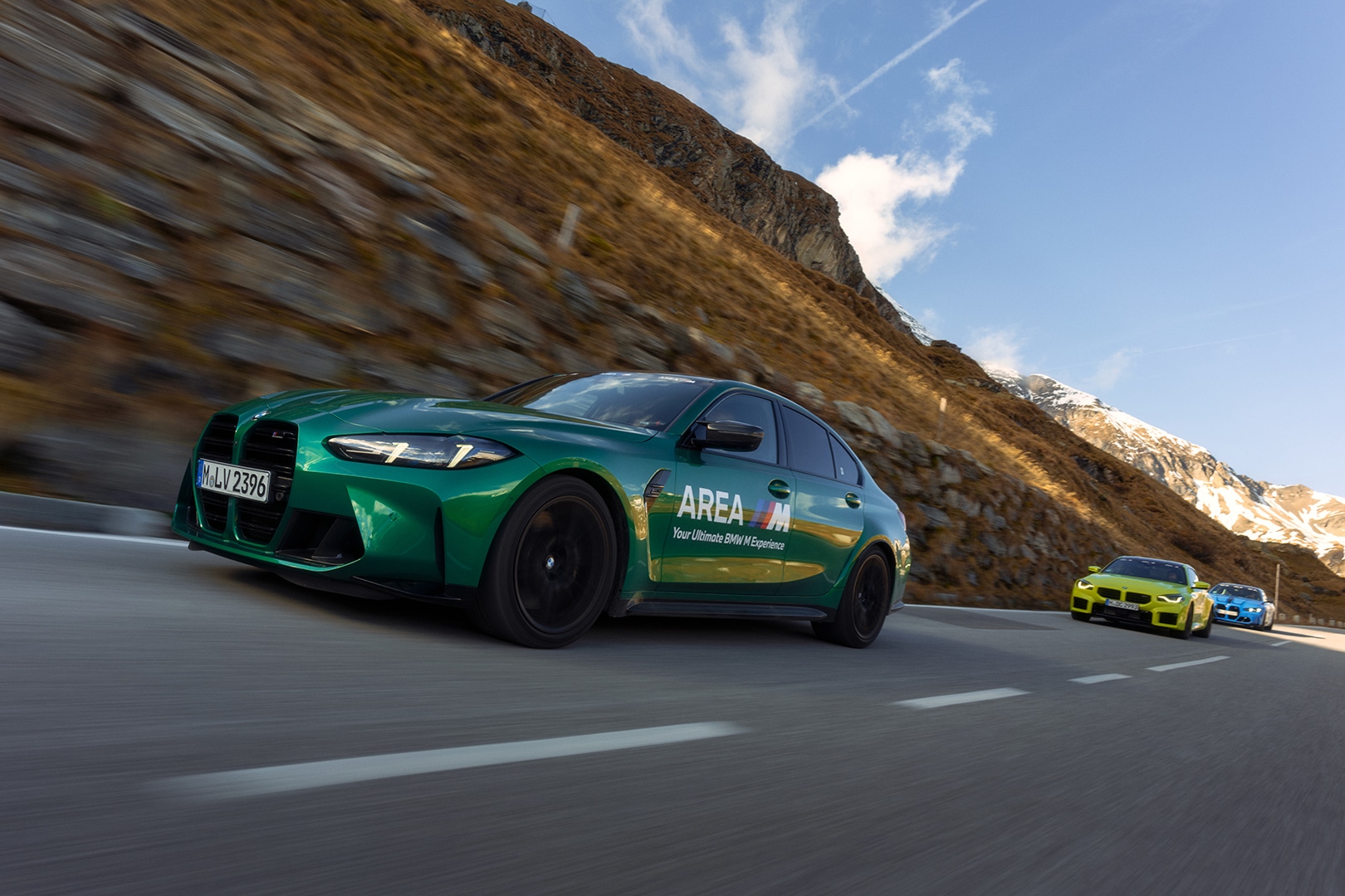 Ein metallisch grüner BMW mit 'AREA M'-Branding fährt auf einer Bergstraße. Dahinter folgen ein gelber und ein blauer BMW. Die Fahrzeuge fahren auf einer kurvenreichen Straße mit felsiger Berglandschaft und blauem Himmel im Hintergrund.