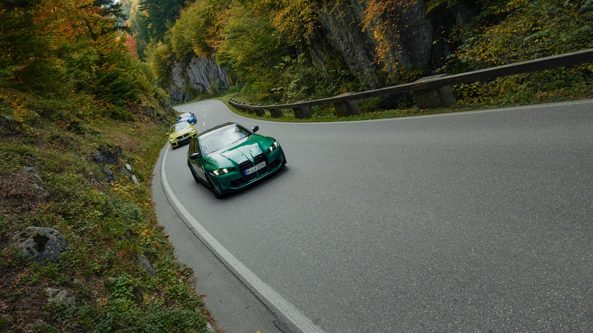 Ein grüner Sportwagen gefolgt von einem gelben Fahrzeug auf einer kurvenreichen Bergstraße, umgeben von herbstlichem Laub. Die geschwungene Asphaltstraße wird von felsigen Klippen und Bäumen mit herbstlichen Farben in Orange, Gelb und Grün gesäumt.