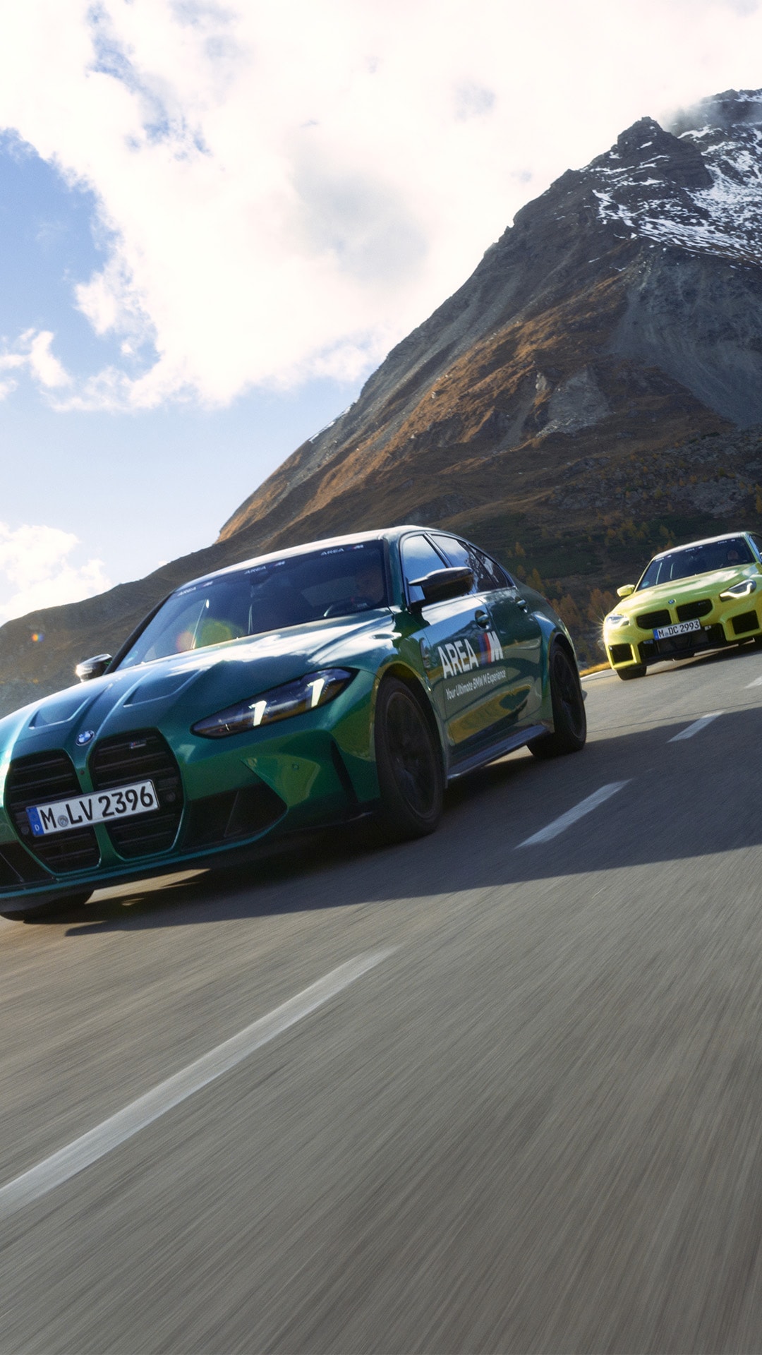 Zwei BMW Sportwagen fahren auf einer Bergstraße. Ein grüner BMW mit 'AREA M'-Branding führt, gefolgt von einem gelben BMW. Sie fahren auf einer kurvenreichen Straße mit einer dramatischen Bergkulisse im Hintergrund, die schneebedeckte Gipfel und einen teilweise bewölkten Himmel zeigt.