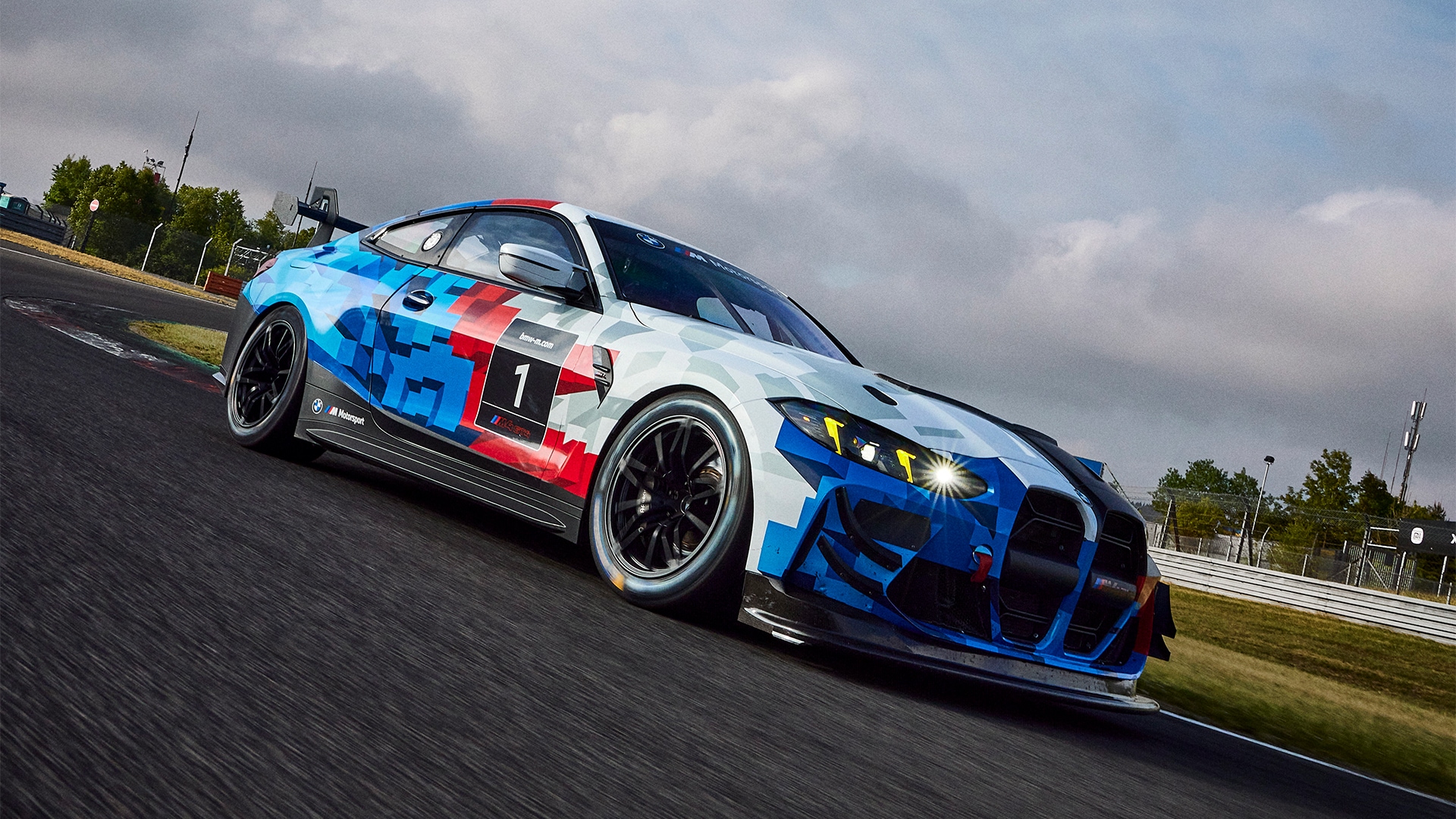 Ein BMW Rennwagen mit der Nummer 1 an der Seite, der eine markante blau-rot-weiße Lackierung mit geometrischem Muster aufweist. Das Fahrzeug steht auf einer Rennstrecke unter bewölktem Himmel, mit eingeschalteten Scheinwerfern und sichtbaren Streckenbegrenzungen im Hintergrund.