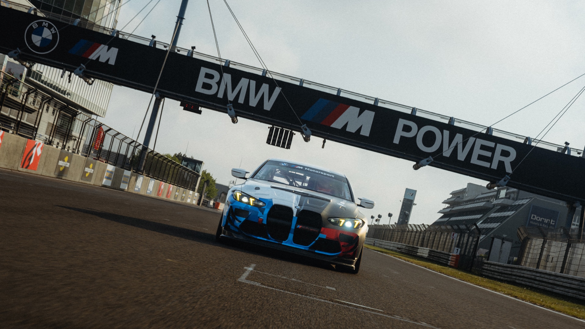 Ein BMW M Rennwagen mit blauen und roten Akzenten fährt auf einer Rennstrecke unter einer großen schwarzen Brücke mit BMW M Power Branding. Die Strecke ist mit Sicherheitsbarrieren und Renninfrastruktur ausgestattet, im Hintergrund ist eine Zuschauertribüne zu sehen.