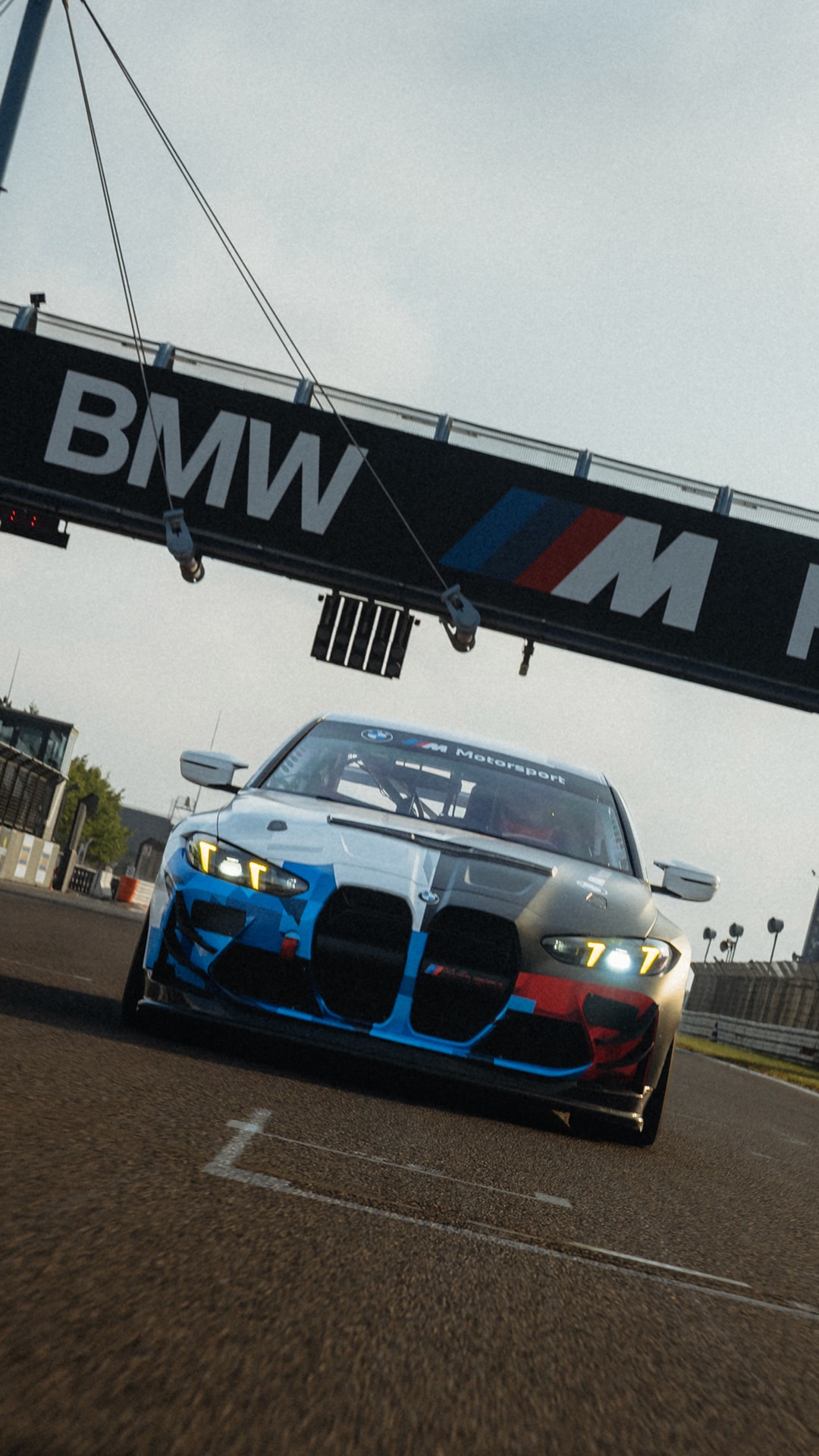 Ein BMW M Motorsport Rennwagen auf einer Rennstrecke, positioniert unter einer großen BMW-Anzeigetafel. Das Fahrzeug hat eine markante blau-weiß-rote Lackierung mit deutlichem BMW M Branding.