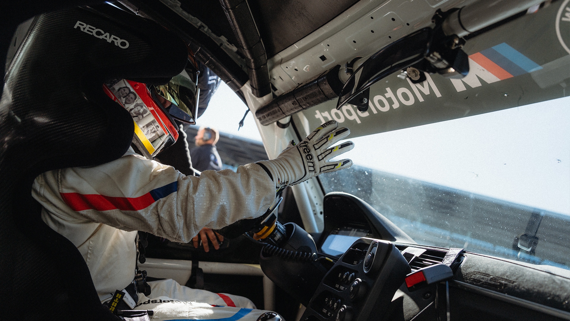 Innenansicht eines Rennwagen-Cockpits mit einem Fahrer in weißem Rennanzug mit roten und blauen Streifen und einem Helm. Der Fahrer trägt Rennhandschuhe und sitzt in einem Recaro-Rennsitz. BMW Motorsport-Branding ist im Innenraum sichtbar.