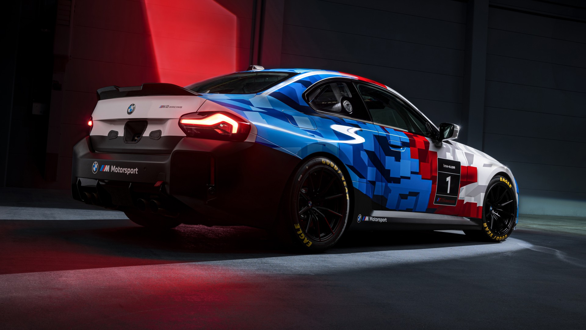 Ein BMW M Motorsport Rennwagen mit einer markanten pixelierten Lackierung in blau, rot und weiß. Das Fahrzeug verfügt über Rennmodifikationen wie einen Heckspoiler und Rennreifen. Es wird in einer dramatischen Umgebung mit roten Lichtakzenten vor einem dunklen Hintergrund präsentiert.