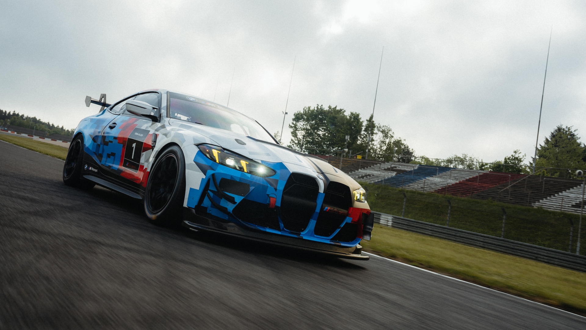 Ein BMW Rennwagen mit einer markanten blau-rot-weißen Tarnlackierung, der auf einer Rennstrecke fährt. Das Hochleistungsfahrzeug verfügt über ein aerodynamisches Karosseriekit mit einem markanten Frontgrill und wird in Bewegung auf der Asphaltbahn aufgenommen, wobei im Hintergrund Tribünen unter einem bewölkten Himmel zu sehen sind.