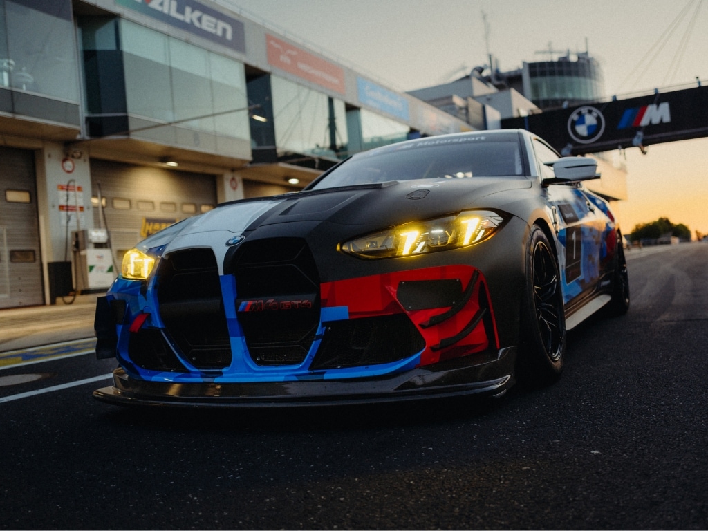 Ein BMW M-Sportwagen mit individueller Rennlackierung in Schwarz, Blau und Rot, der auf einer Rennstrecke steht. Das Fahrzeug hat markante gelbe Scheinwerfer und befindet sich vor Boxengaragen mit BMW und anderen Motorsport-Markierungen im Hintergrund während des Sonnenuntergangs.
