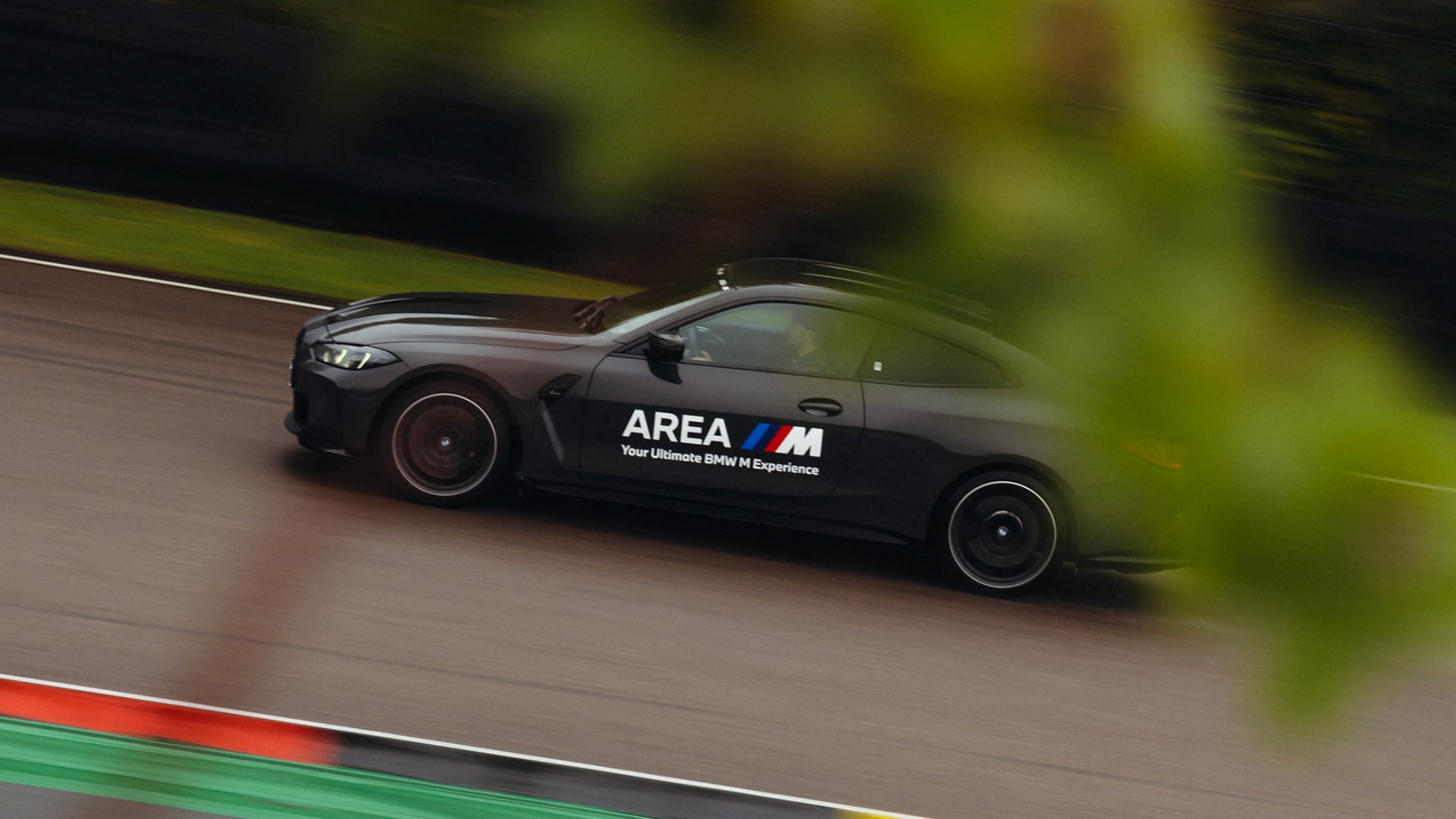 A black BMW M series sports car in motion on a race track. The vehicle displays 'AREA M' branding with the BMW M logo on its side and the tagline 'Your Ultimate BMW M Experience'. The car is captured with motion blur against a blurred green background, suggesting high speed.