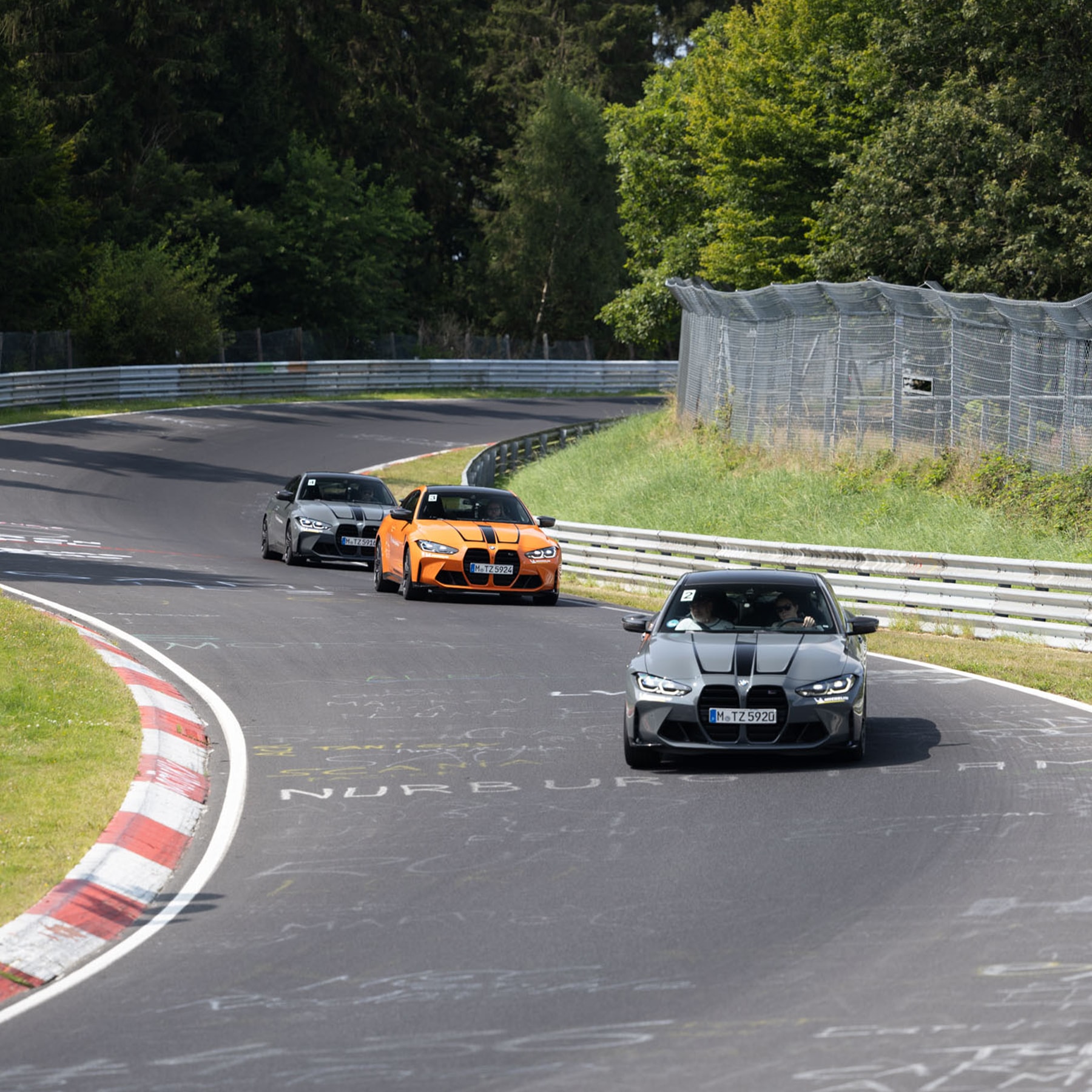 Three BMW sports cars driving on the Nürburgring race track. A gray BMW leads, followed by an orange BMW and another gray BMW. The cars are navigating a curve on the famous track, surrounded by safety barriers and green forest.