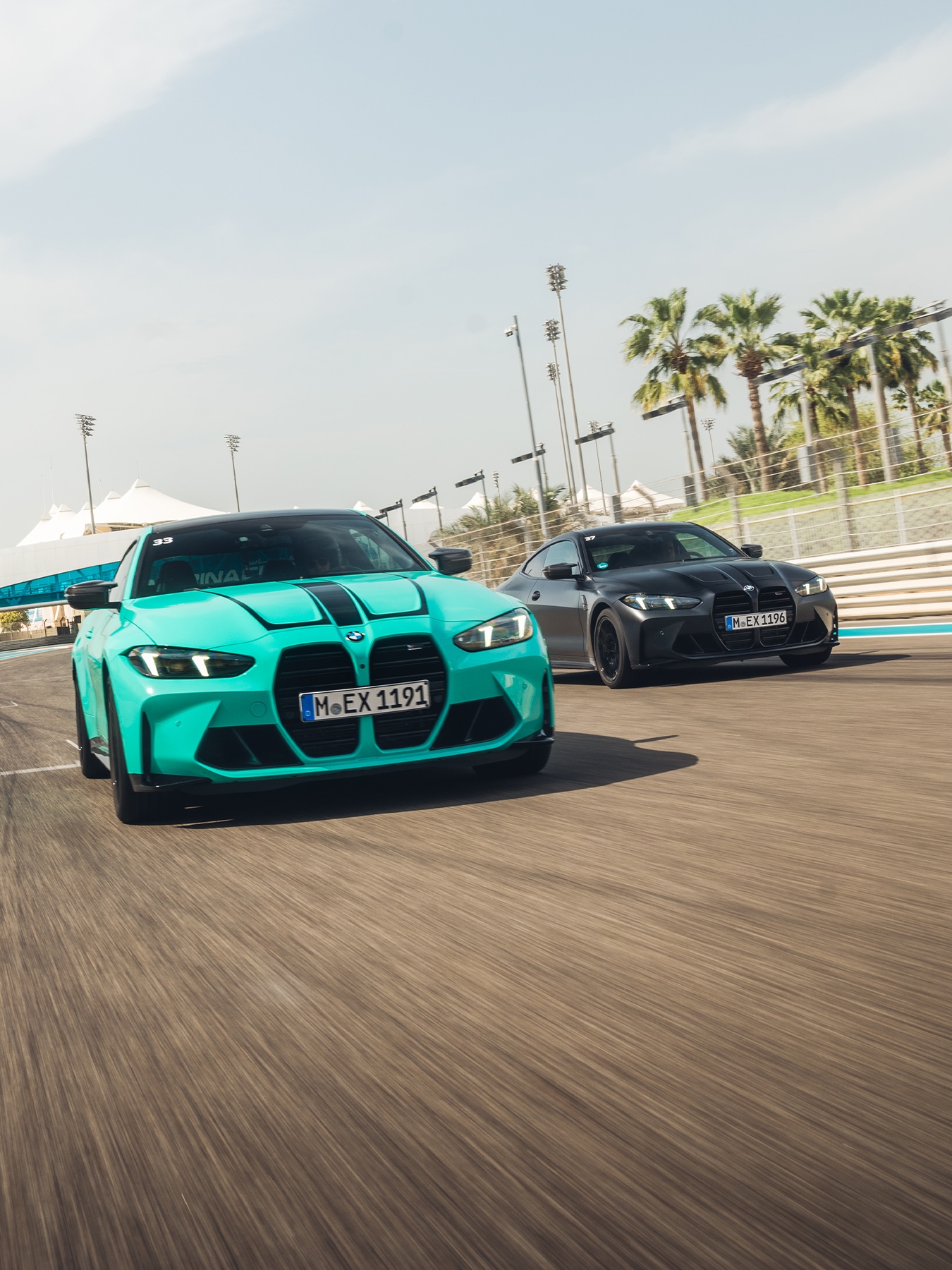 Two BMW sports cars driving on a racetrack, with a turquoise model in front and a black model behind. Palm trees and stadium lights are visible in the background under a clear blue sky.