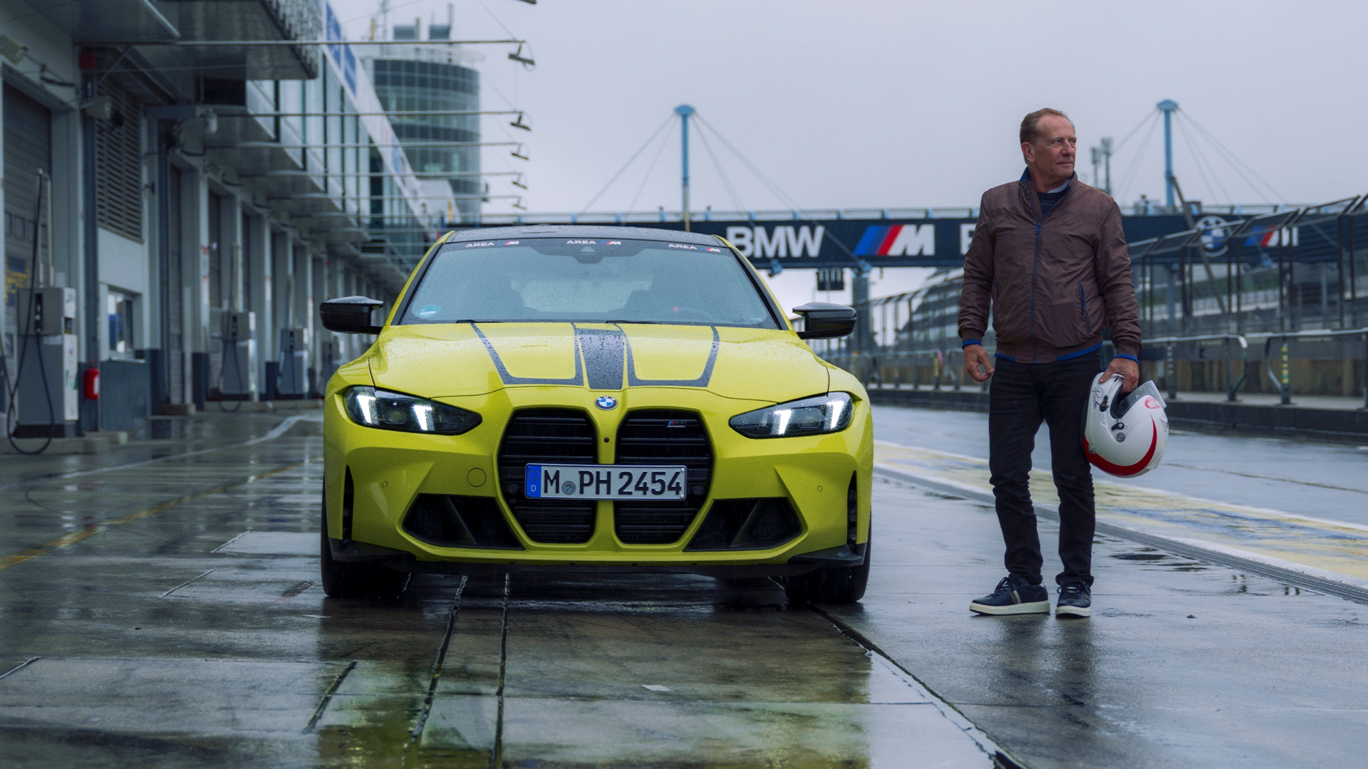 A bright yellow BMW M sports car parked on a wet racetrack pit lane. A person in a brown jacket stands beside the car holding a white helmet. In the background, there's a BMW M branded bridge spanning the track on a cloudy day.