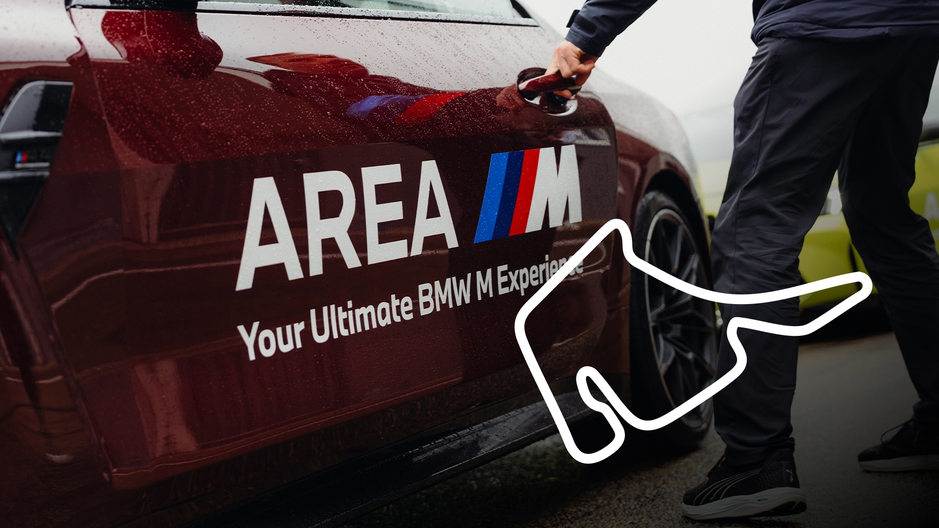 Ein dunkelrotes BMW-Fahrzeug mit "AREA M"-Branding und dem Text "Your Ultimate BMW M Experience" auf der Seite. Eine Person in dunkler Kleidung öffnet die Autotür. Im unteren rechten Bereich des Bildes ist ein weißer Umriss einer Rennstrecke zu sehen.
