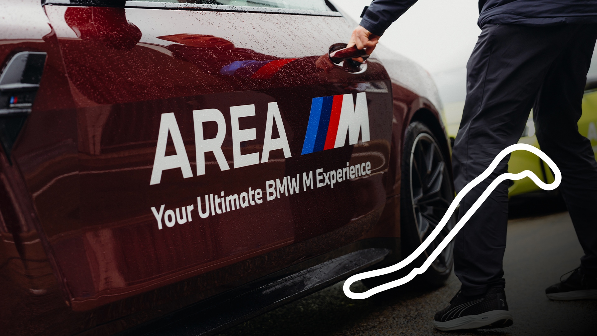 Ein dunkelroter BMW mit "AREA M"-Branding und dem Text "Your Ultimate BMW M Experience" auf der Tür. Eine Person in dunkler Kleidung öffnet die Autotür. Das Fahrzeug scheint mit Regentropfen bedeckt zu sein, und in der rechten unteren Ecke des Bildes ist ein weißer Umriss einer Rennstrecke zu sehen.