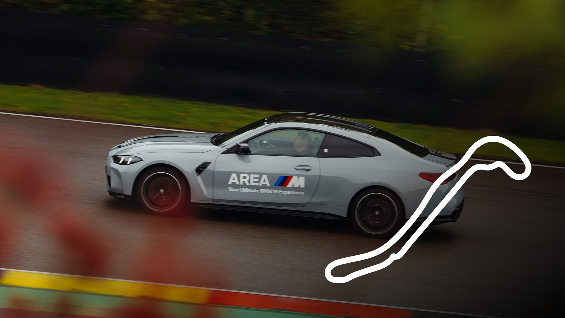 Ein hellblaues BMW M Fahrzeug in Bewegung auf einer Rennstrecke. Das Fahrzeug trägt die Aufschrift 'AREA M' mit dem BMW M-Logo an der Seite. Eine weiße Umrisslinie der Streckenkurve ist in der unteren rechten Ecke des Bildes sichtbar.
