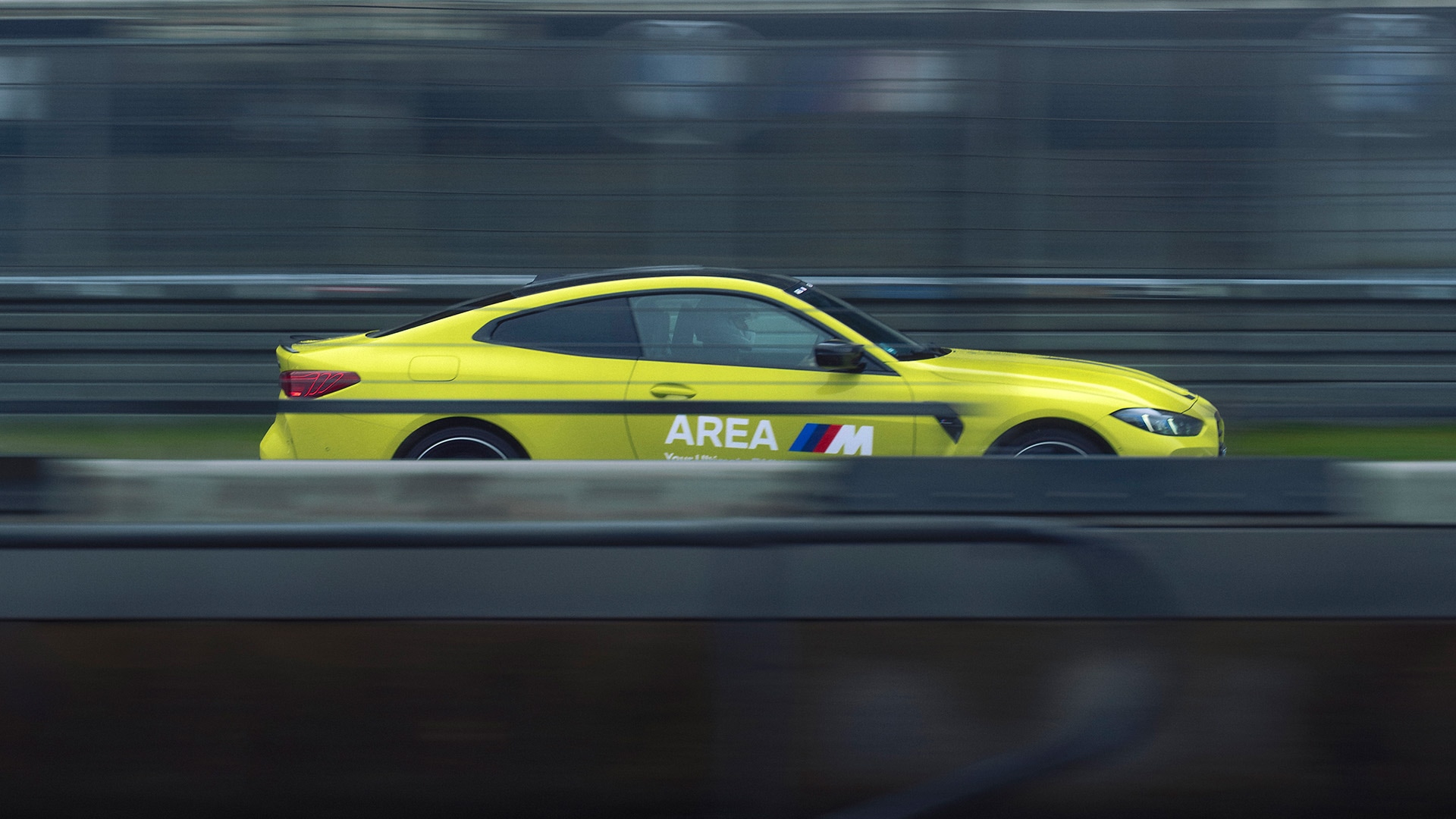 Ein leuchtend gelber BMW Sportwagen mit 'AREA M' Beschriftung und den charakteristischen BMW M-Farben an der Seite, aufgenommen in Bewegung auf einer Rennstrecke mit verschwommenem Hintergrund, was auf hohe Geschwindigkeit hindeutet.